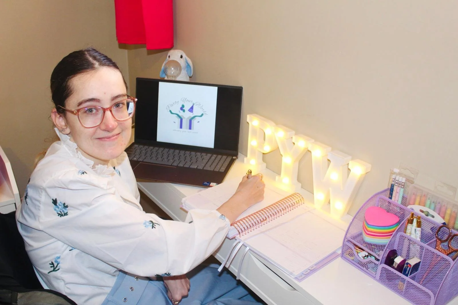 Young woman with glasses sitting at a desk, working on a notebook, surrounded by colourful stationery, a laptop displaying a logo, and illuminated decorative letters spelling 'PYW' on the desk.