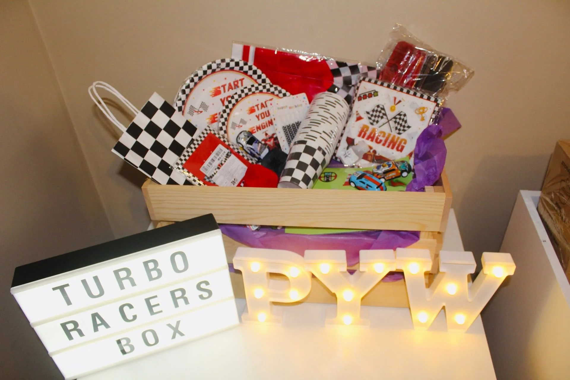A wooden box filled with race car-themed party supplies, including checkered flag napkins, cups, and decorations. A lightbox sign reads 'TURBO RACERS BOX,' and a decorative marquee sign spells out 'PYW' with bright bulbs.