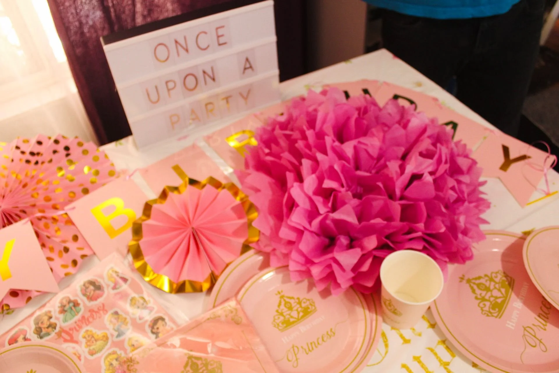 Pink and gold princess-themed party decorations, including pink tissue paper pom-pom, paper fans, plates, cups, and a letter board that says 'Once Upon a Party'.