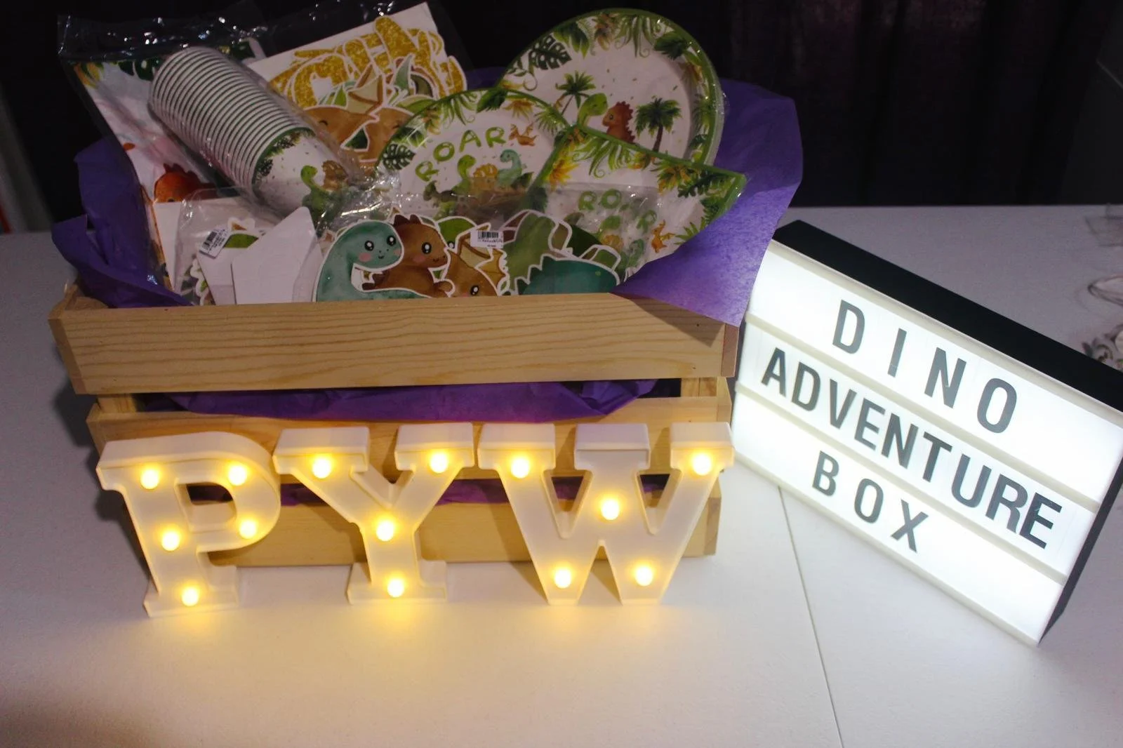 A box filled with dinosaur-themed paper plates and cups, decorated with colorful dinosaurs and jungle scenery. In front of the box is a light-up sign spelling 'PYW'. To the right, there is a lightbox displaying 'DINO ADVENTURE BOX' on a white surface.
