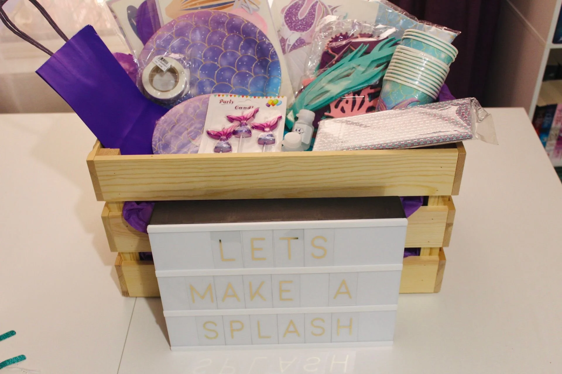 A wooden crate filled with party supplies such as paper plates, napkins, cups, and decorations. In front, a lightbox displays the words 'LET'S MAKE A SPLASH' in Gold letters.