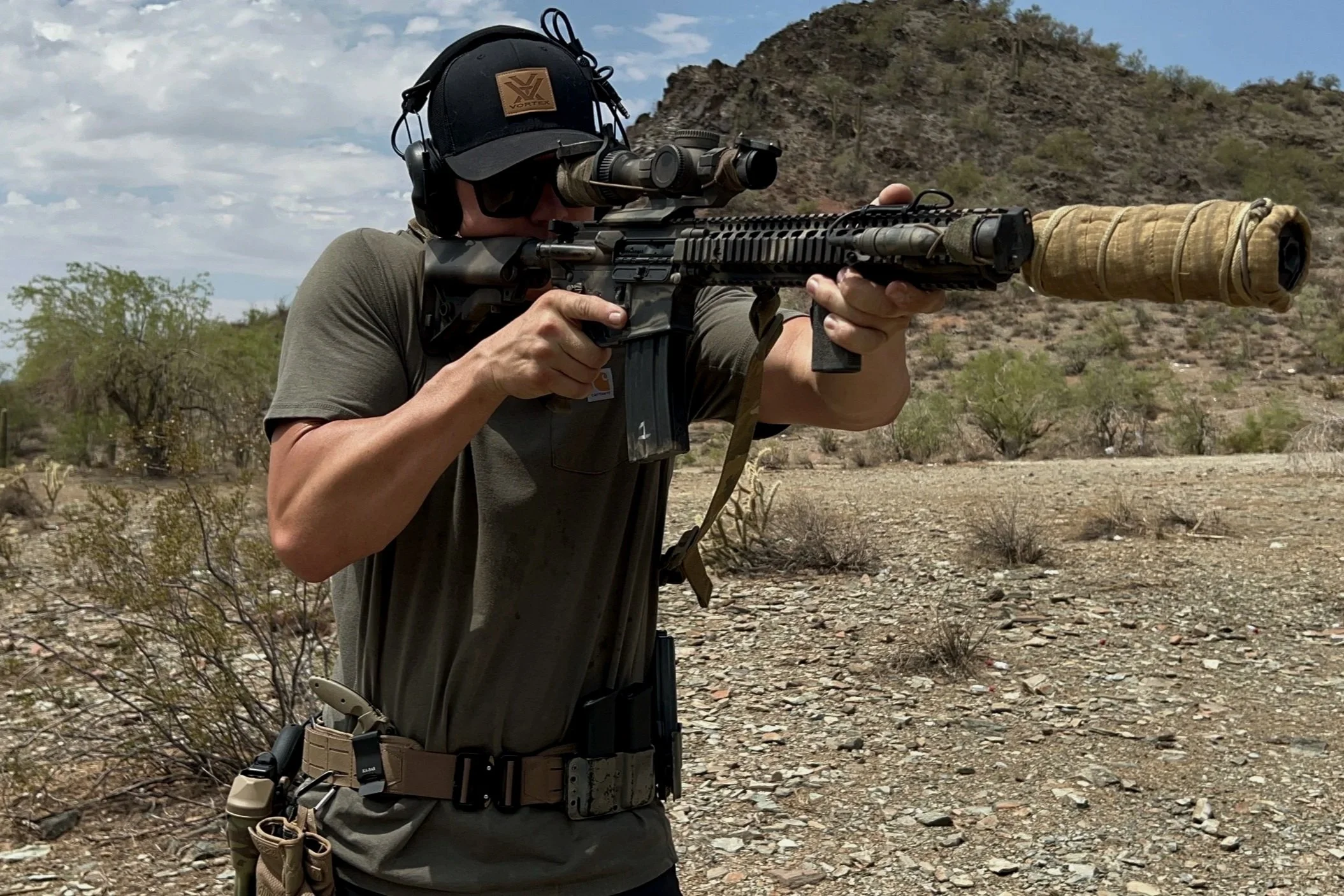 A man wearing tactical gear aiming a scoped rifle in a desert landscape.