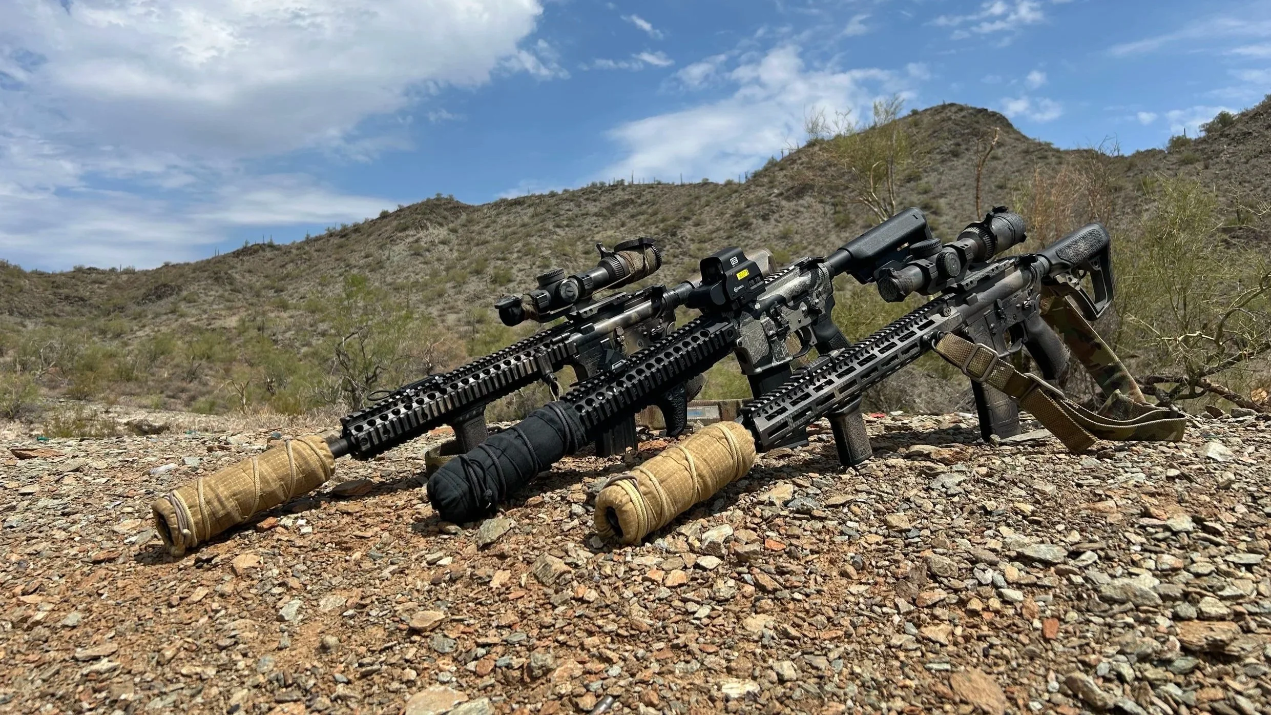 Two tactical rifles lying on rocky ground in a desert landscape with mountains and sparse vegetation under a partly cloudy sky.