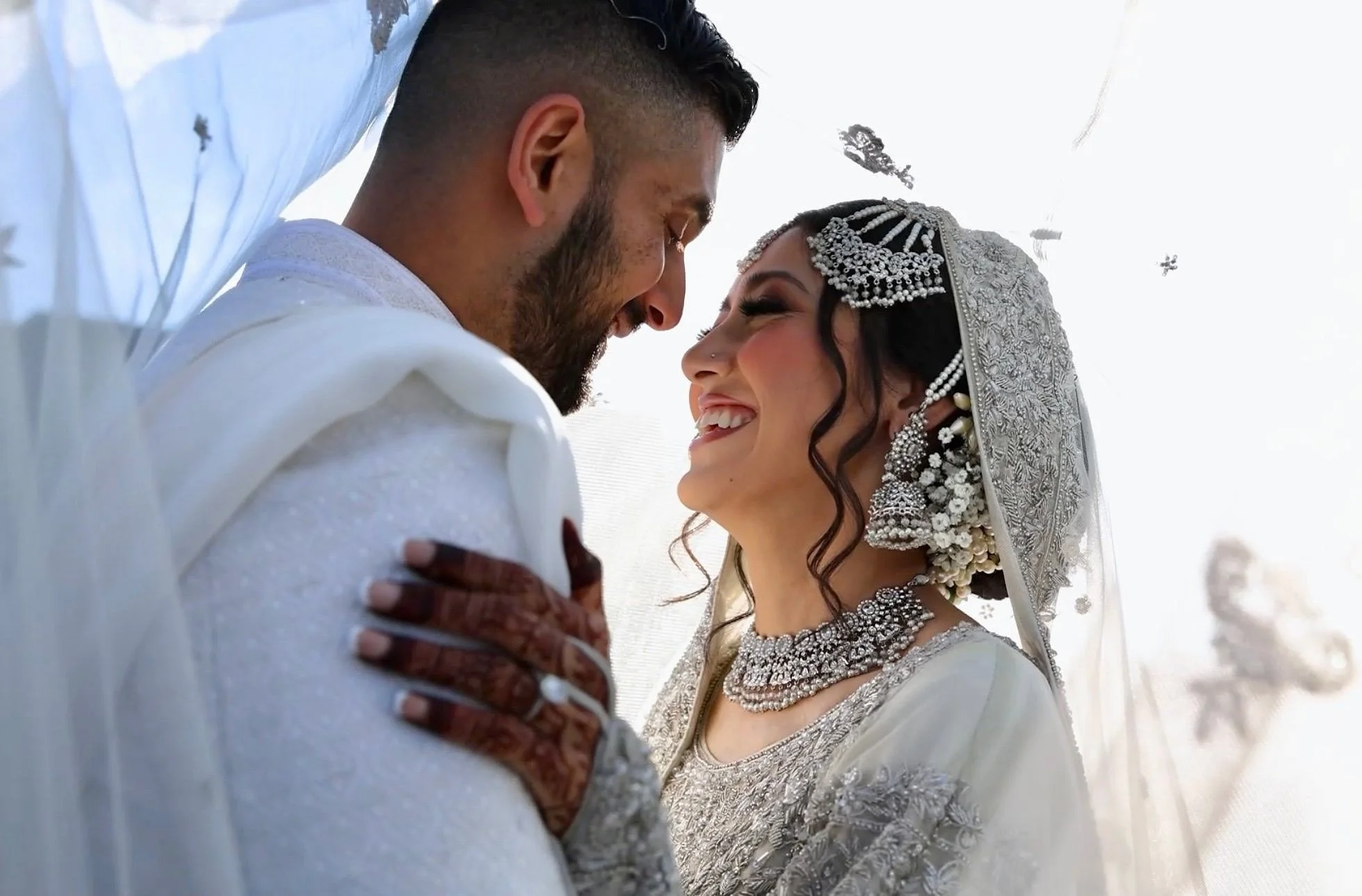 A bride and groom smiling and touching foreheads during their wedding, dressed in traditional wedding attire with jewelry.