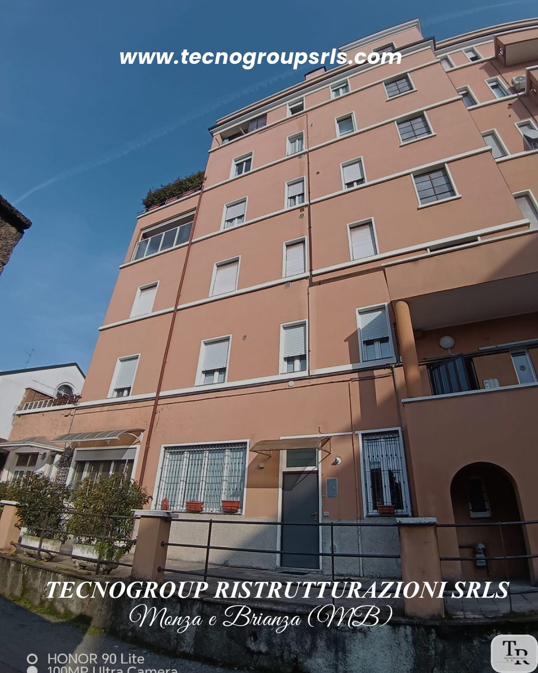 Photograph of a multi-story pink residential building with white window shutters, some open and some closed, and a small balcony with plants and a railing. The sky is blue with a few wispy clouds. Text overlays in white and black read "www.tecnogroup