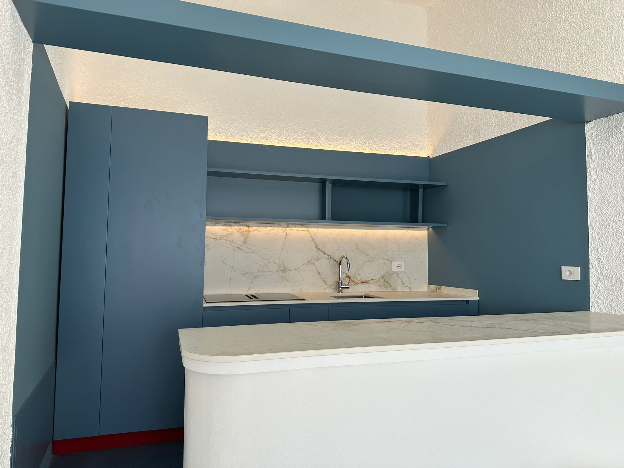 Modern kitchen with blue cabinetry, marble backsplash, and a built-in sink with a faucet. Open shelving above the sink and a white curved countertop in the foreground.