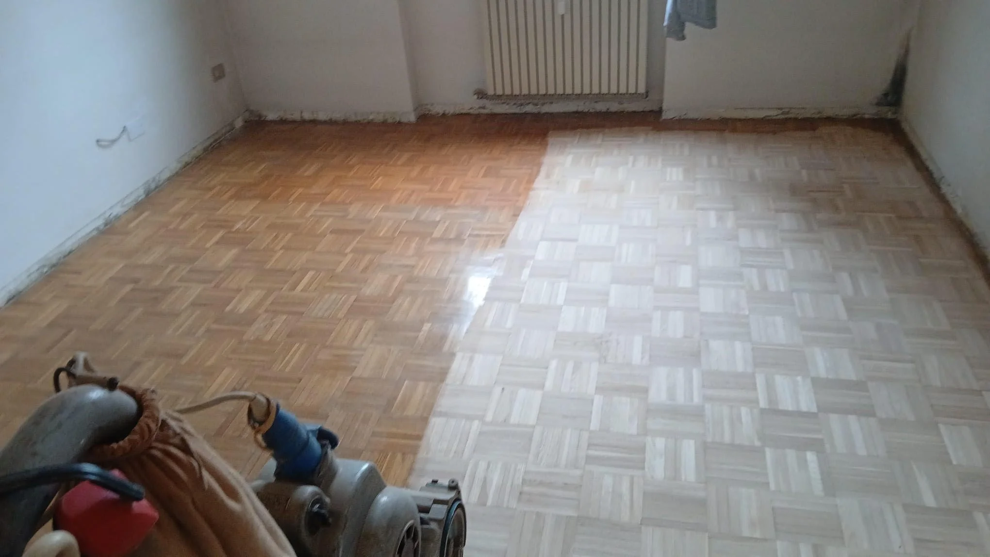 A room with partially renovated parquet flooring, with some areas having a darker finish and others lighter. There is a vacuum cleaner and some cleaning supplies in the lower left corner. The walls are white, with an electrical outlet on the left wall and a window or radiator at the back.