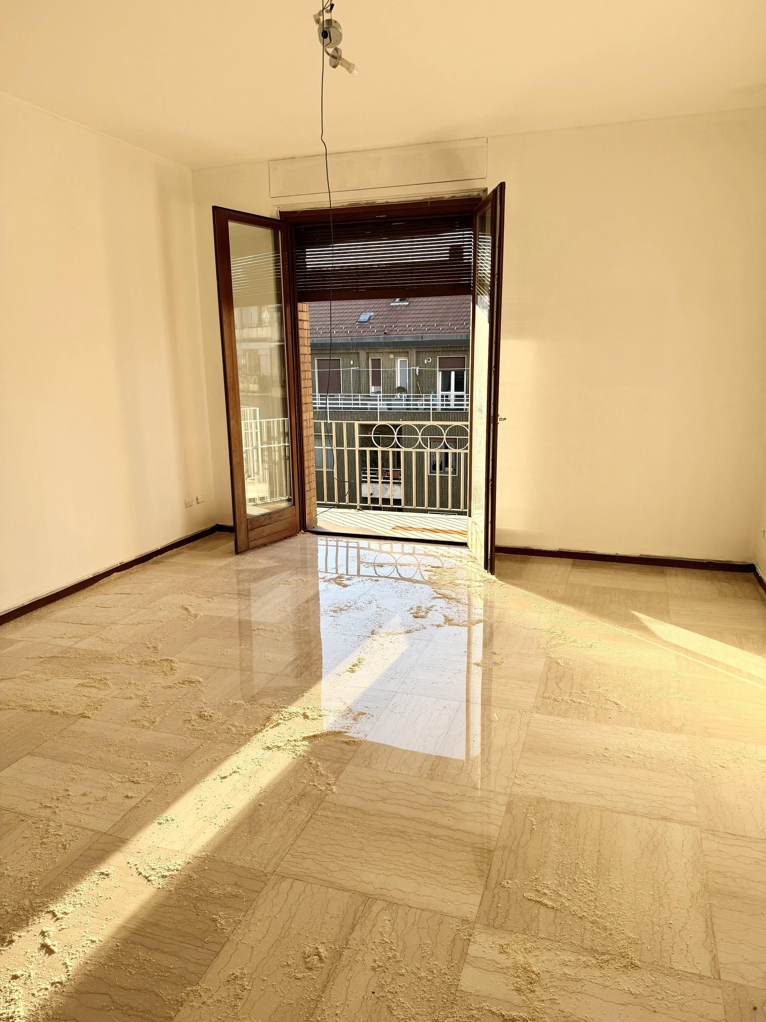 Empty room with open balcony door, sunlight streaming in, tiled floor, and a balcony railing visible outside.