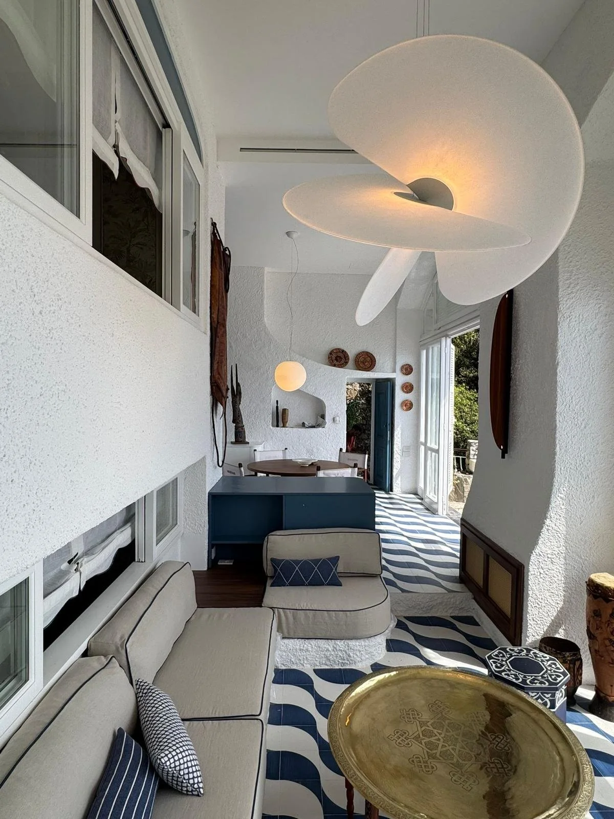 Interior living room with white textured walls and ceiling, modern abstract ceiling lamps, beige sectional sofa with striped and dotted throw pillows, blue and white patterned tiled floor, round gold coffee table, and open balcony door revealing greenery outside.