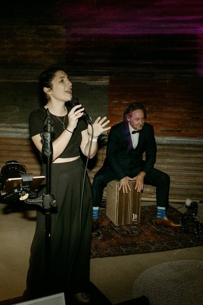 A woman singing into a microphone, wearing a black cropped top and pants, with a man playing a cajón drum behind her, dressed in a black suit and bow tie, in a dimly lit venue with wooden wall background.
