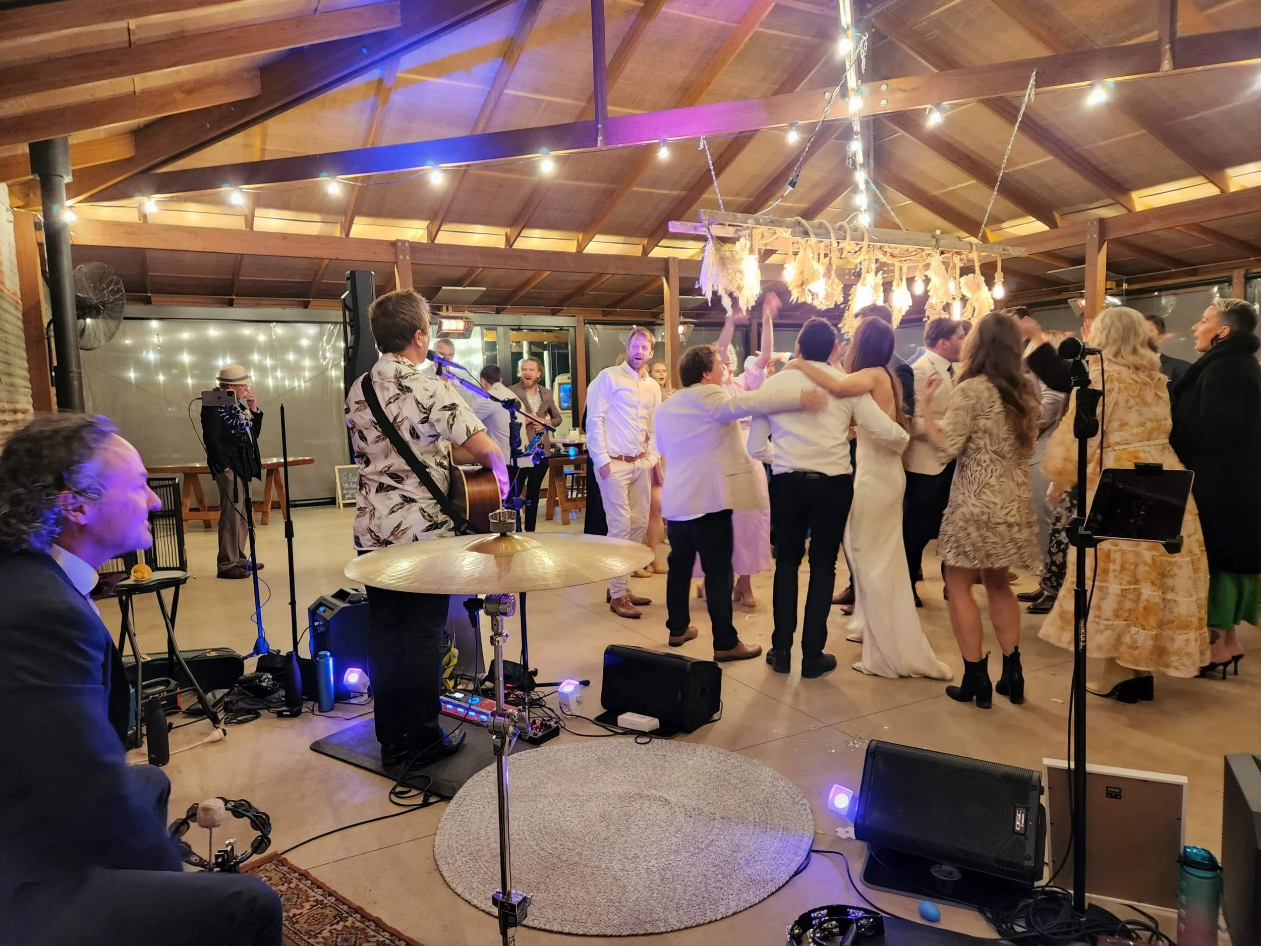 People dancing and celebrating at a wedding reception in a rustic venue with string lights and decorations.