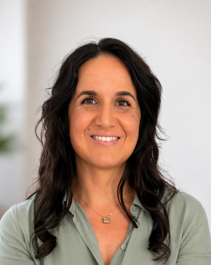 A woman with dark, wavy hair smiling at the camera, wearing a light green blouse and a delicate necklace.