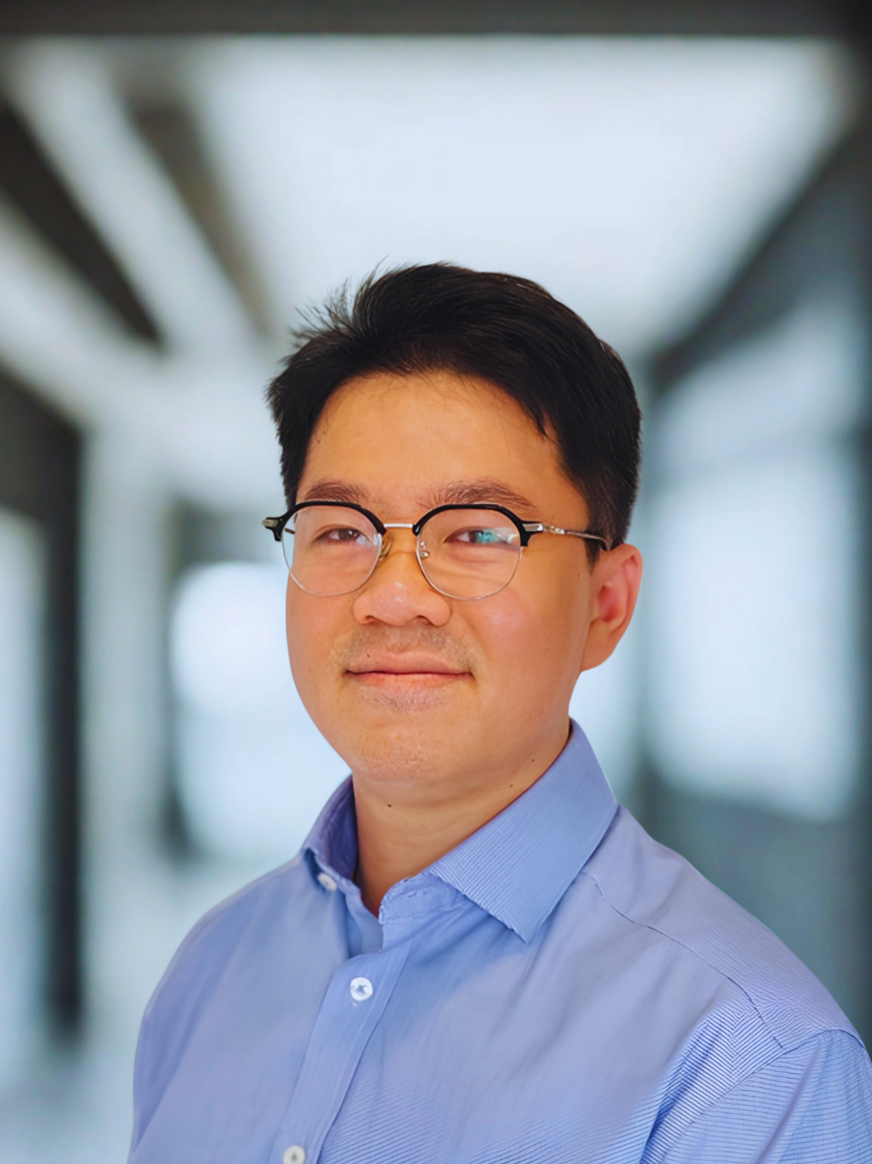 A professional Asian man wearing glasses and a light blue dress shirt, standing in a modern indoor corridor with blurred background.