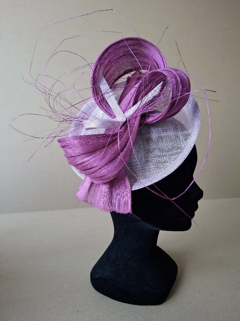 Une coiffe élaborée en rubans de satin violet, blanc et des fils fins, exposée sur un mannequin noir. La coiffe comporte des spirales, des voiles et un design artistique sophistiqué.