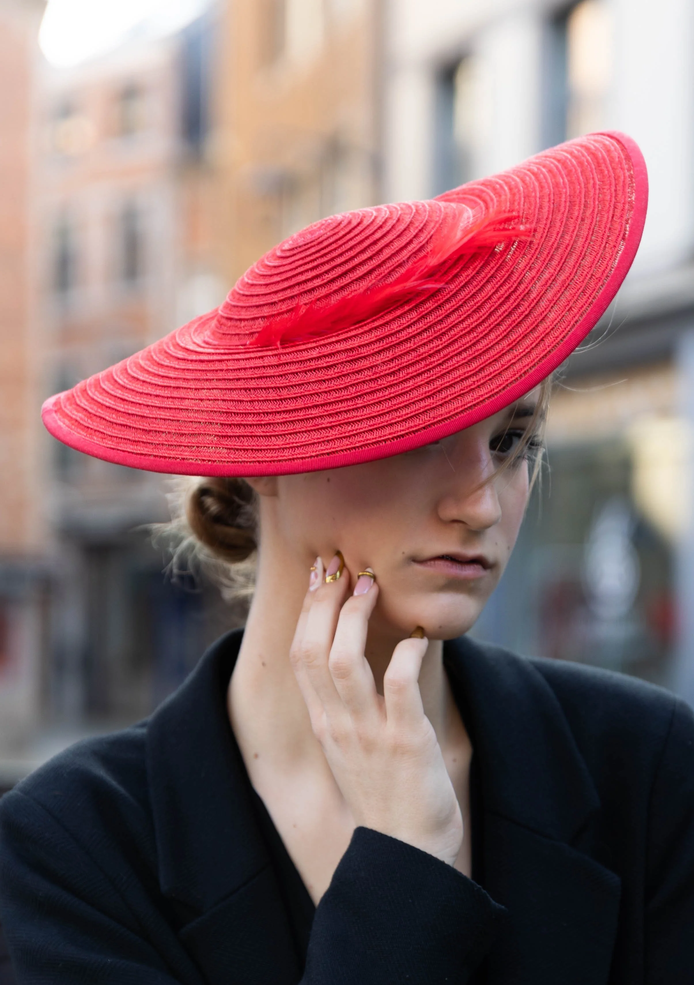 Une femme portant un large chapeau rouge avec un ruban en tissu translucide, dans une zone urbaine, portant une veste noire, avec une main près du visage.