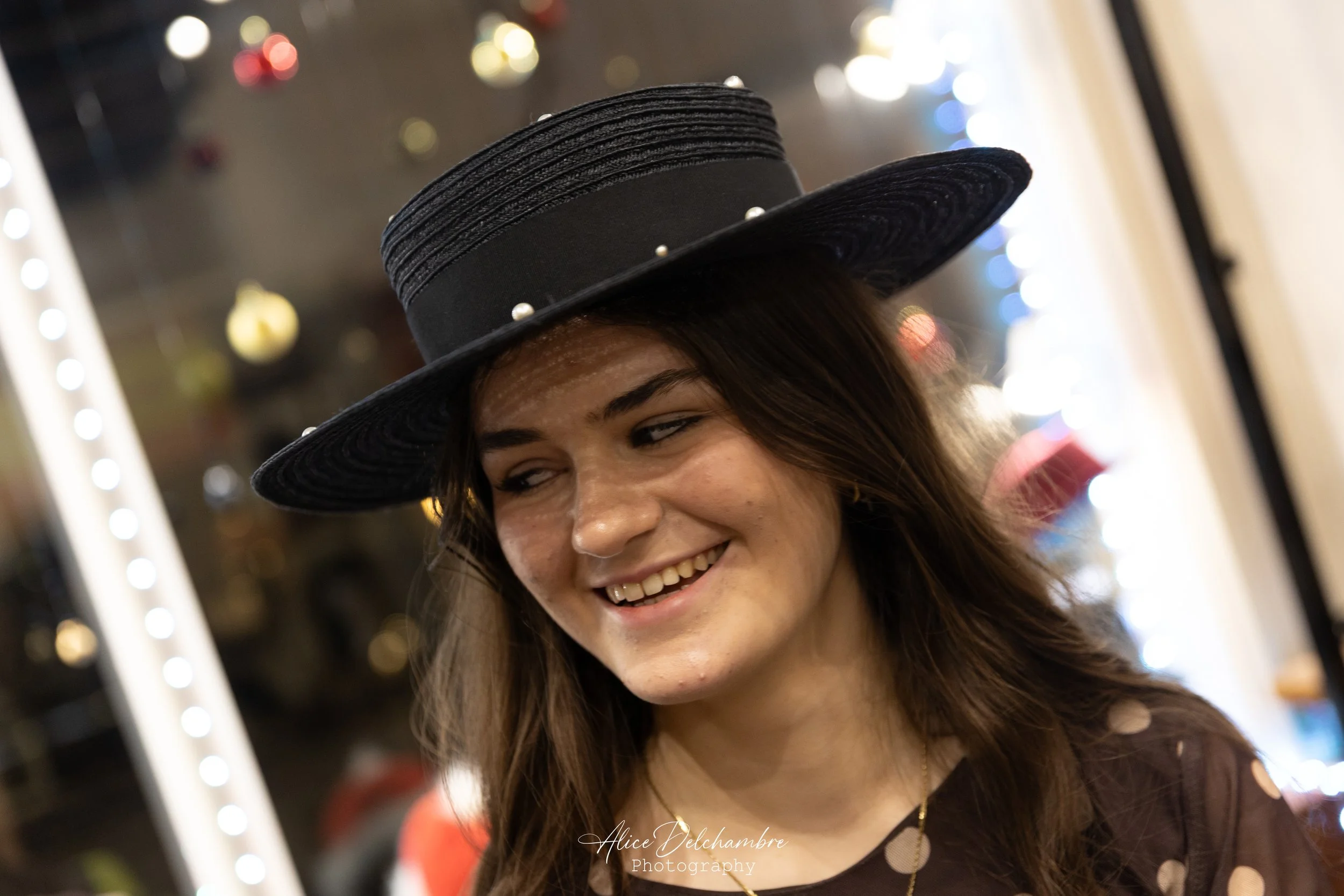 Une femme porte un grand chapeau noir avec des perles et sourit, arrière-plan flou avec des décorations lumineuses.