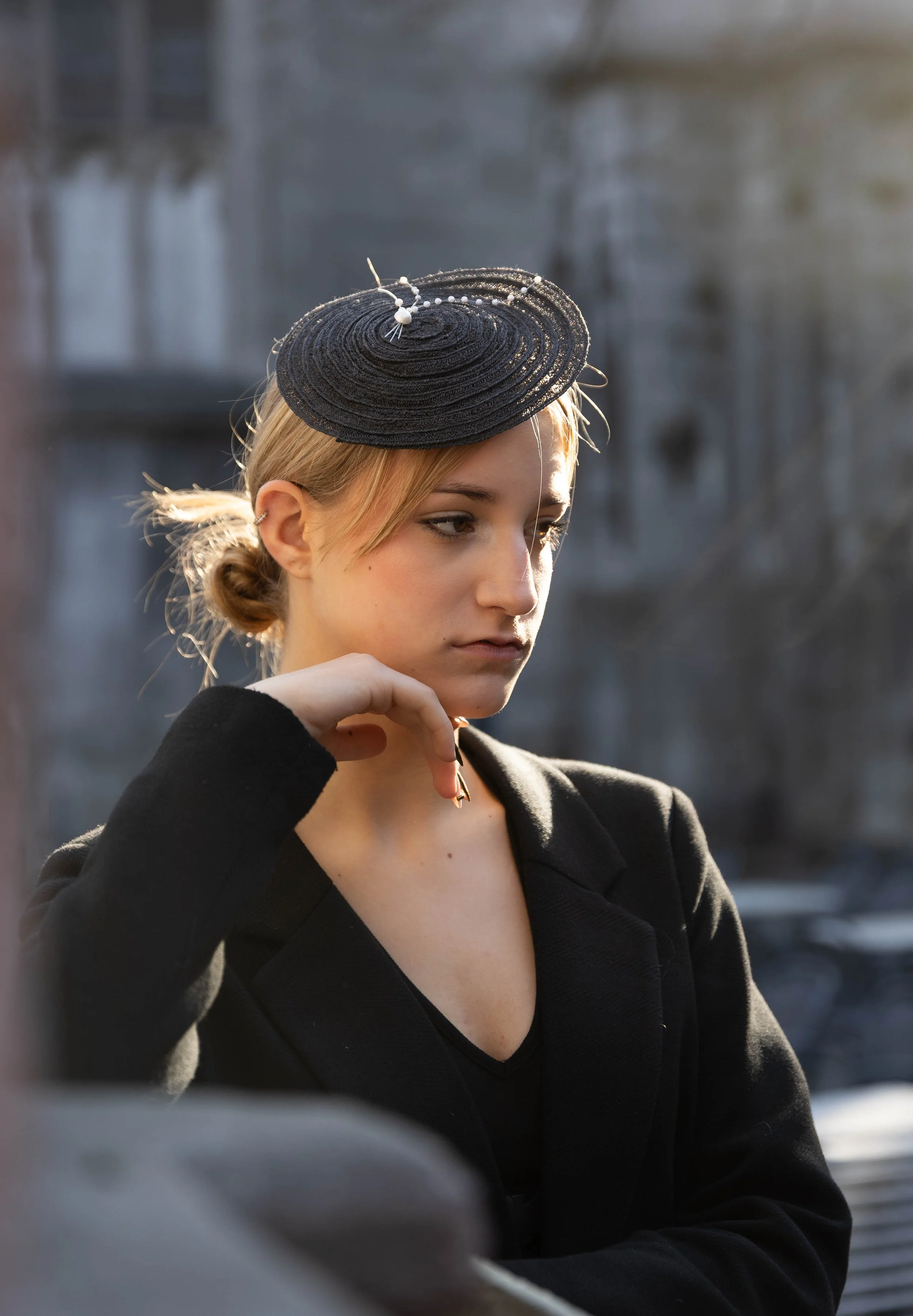 Une femme blonde portant un chapeau noir avec des perles et une veste noire dans une fête, un cocktail regardant pensivement.