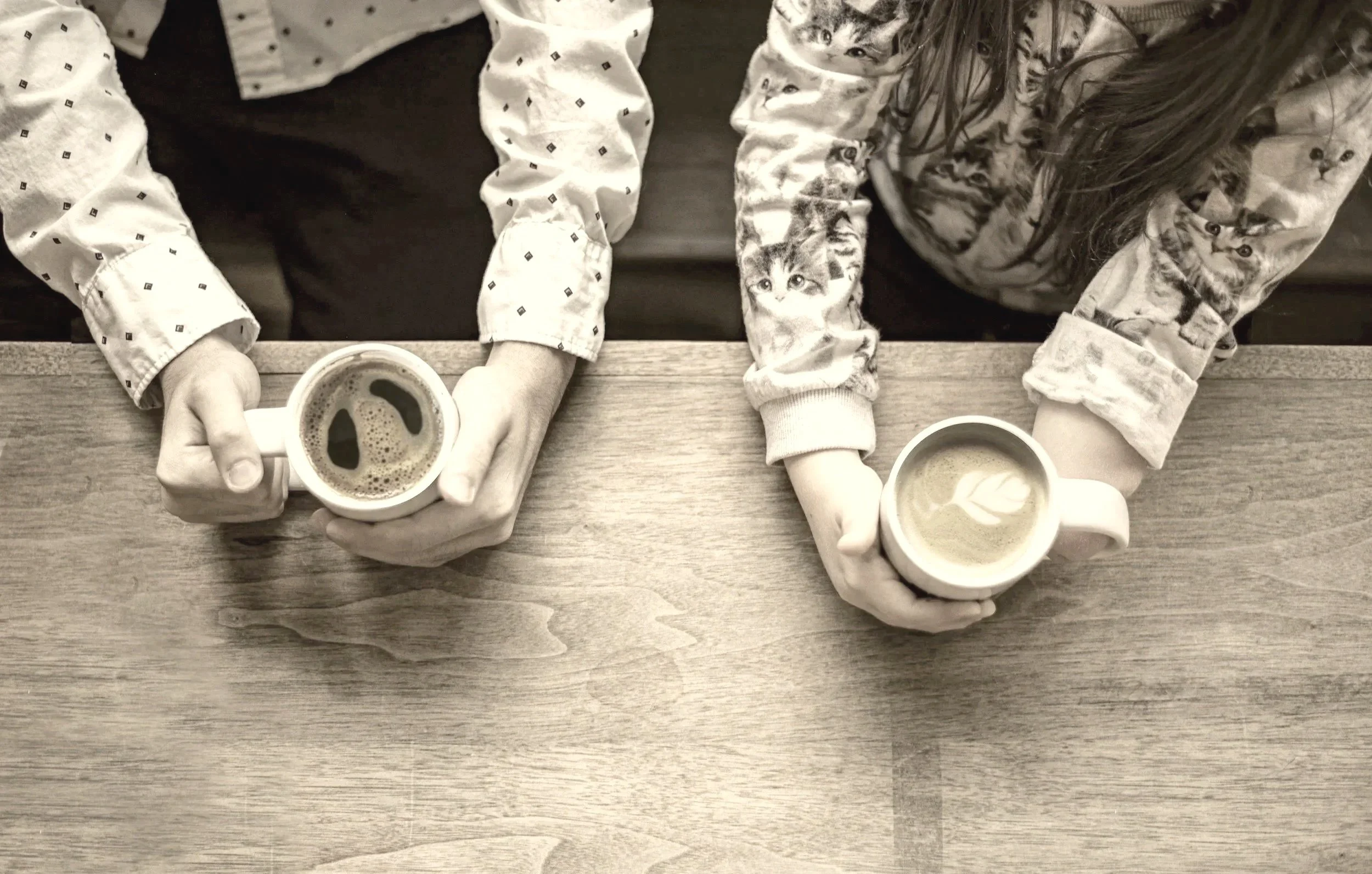 Two people sitting at a wooden table, hands visible each holding muted coffee cups.