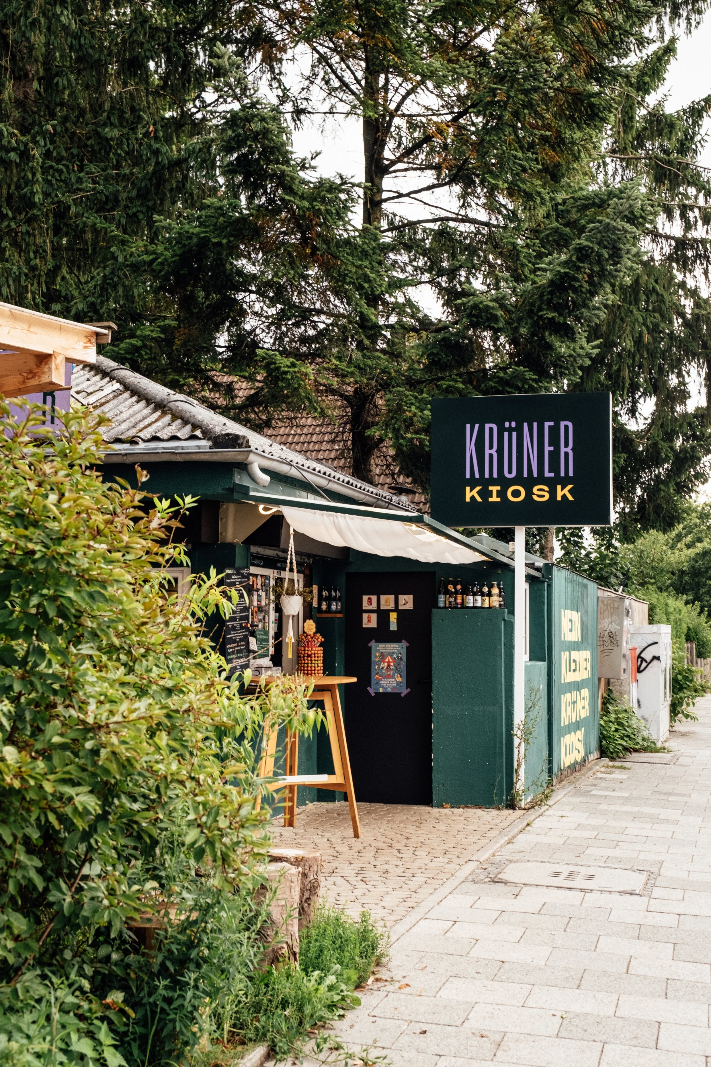 Kleiner Kiosk mit einem schwarzen Schild, auf dem in lila und gelb KRÜNER KIOSK steht, am Straßenrand, umgeben von Büschen und Bäumen, mit einem kleinen Tisch und Getränkeflaschen im Inneren.