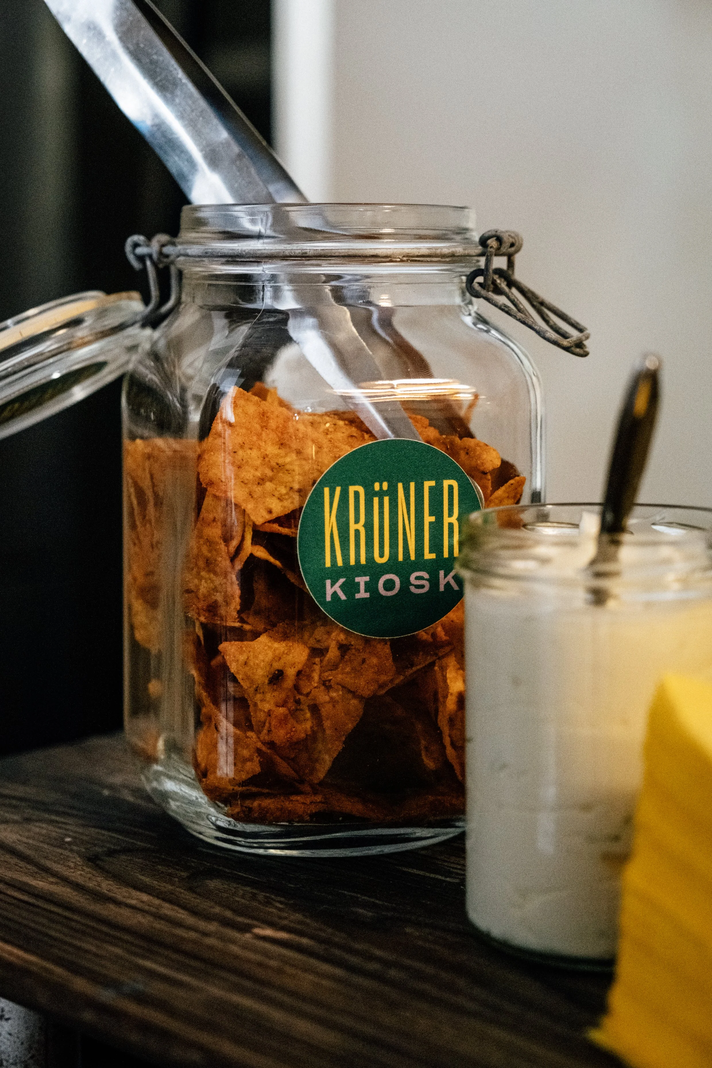 Glas mit Keksen, ein weiterer Behälter mit weißer Creme, kleine Stücke von Avocado, Laden mit einem grünen Aufkleber mit gelber und lilafarbener Schrift, der 'KRÜNER KIOSK' sagt.