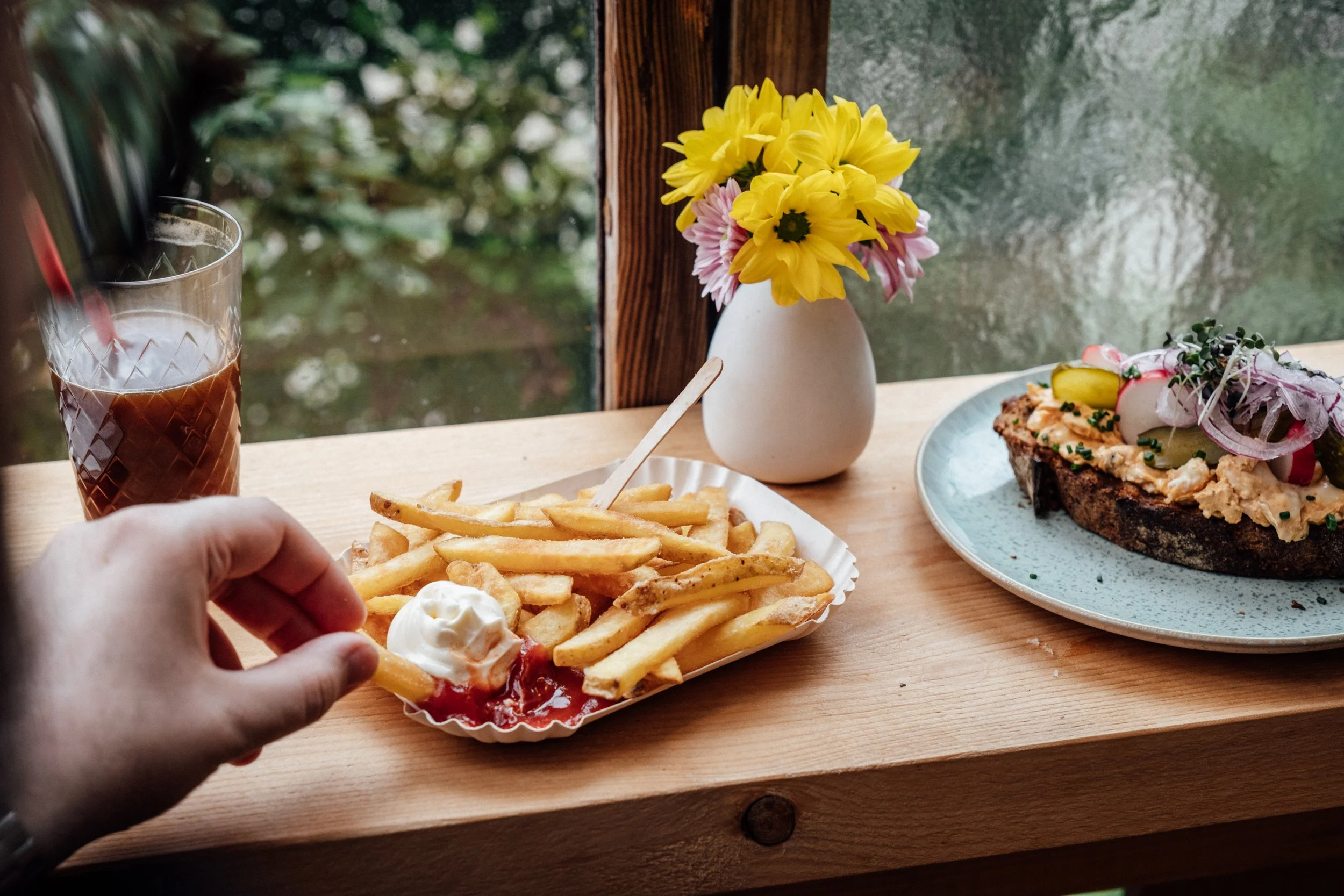 Fries mit Ketchup und Mayonnaise, eine Hand greift nach einer Pommes, ein Glas Cola, eine Vase mit gelben und pinken Blumen, ein Teller mit Aufschnitt und Zwiebeln auf einem Holztisch, Blick durch ein Fenster in die Natur.