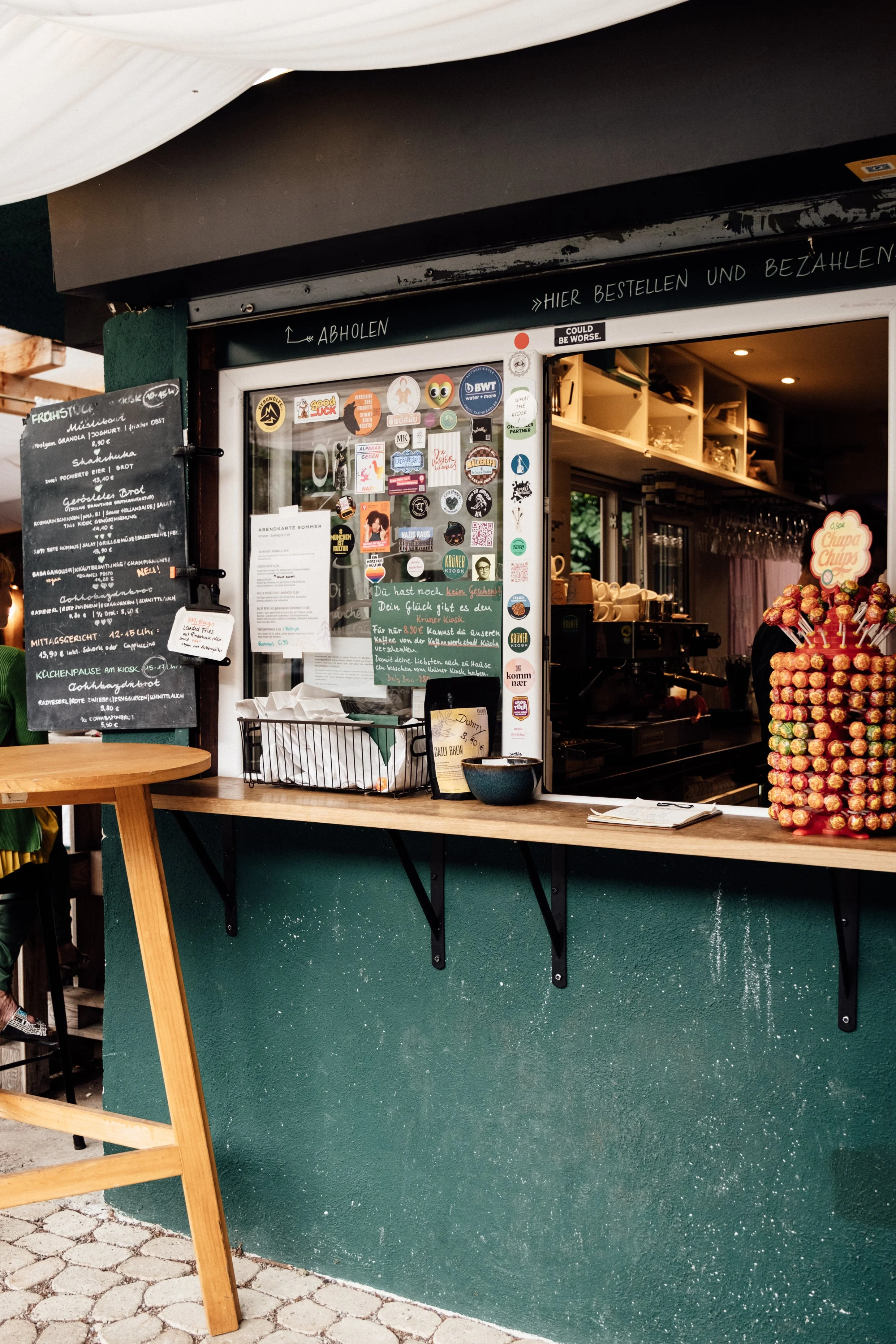 Bestell- und Abholfenster eines Cafés mit zahlreichen Stickern auf der Scheibe, einem schwarzen Brett mit Menü, einem Korb mit Papier, einer Tüte, und einer großen Stange mit Lutschern