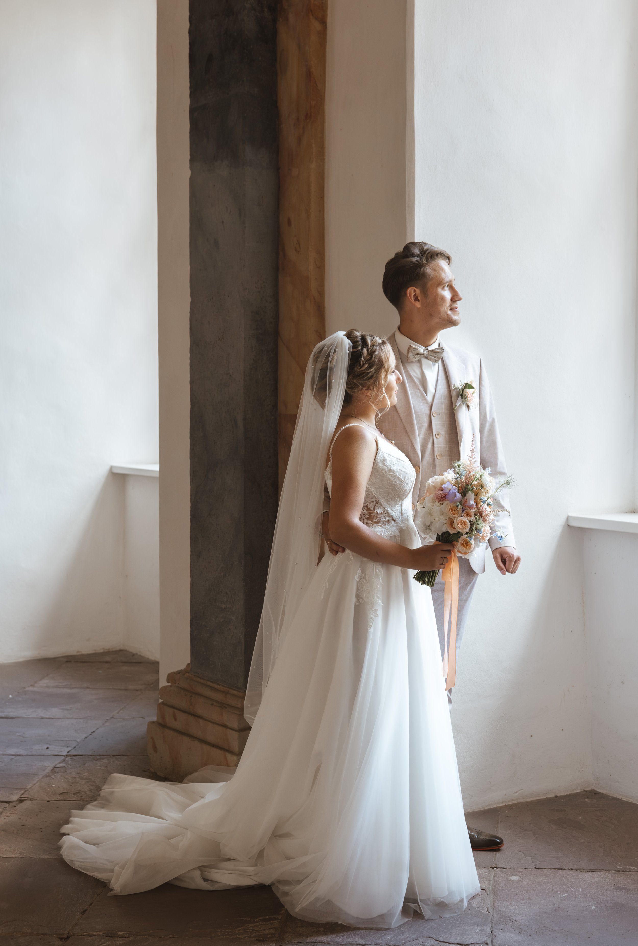 Ein brautpaar steht nebeneinander in einem hellen Raum, beide schauen nach draußen. Die Braut trägt ein weißes Hochzeitskleid mit Schleier und hält einen Blumenstrauß. Der Bräutigam trägt einen hellen Anzug mit Fliege.