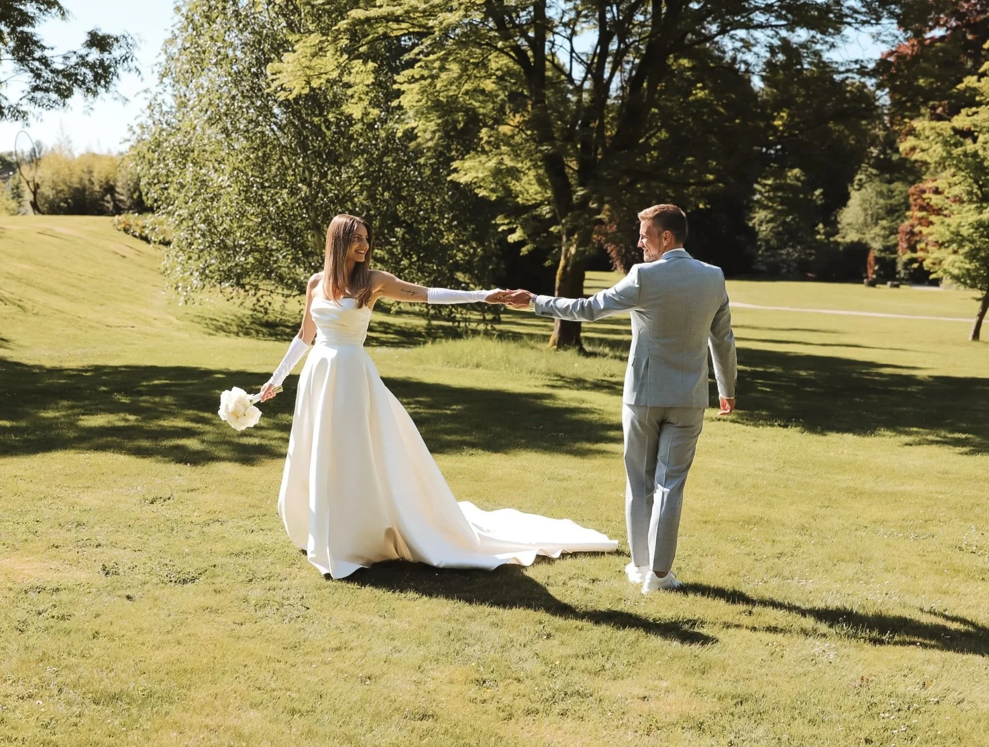 Ein Brautpaar tanzt auf einer grünen Wiese unter Bäumen im Sonnenschein, sie trägt ein weißes Hochzeitskleid und er einen hellgrauen Anzug.