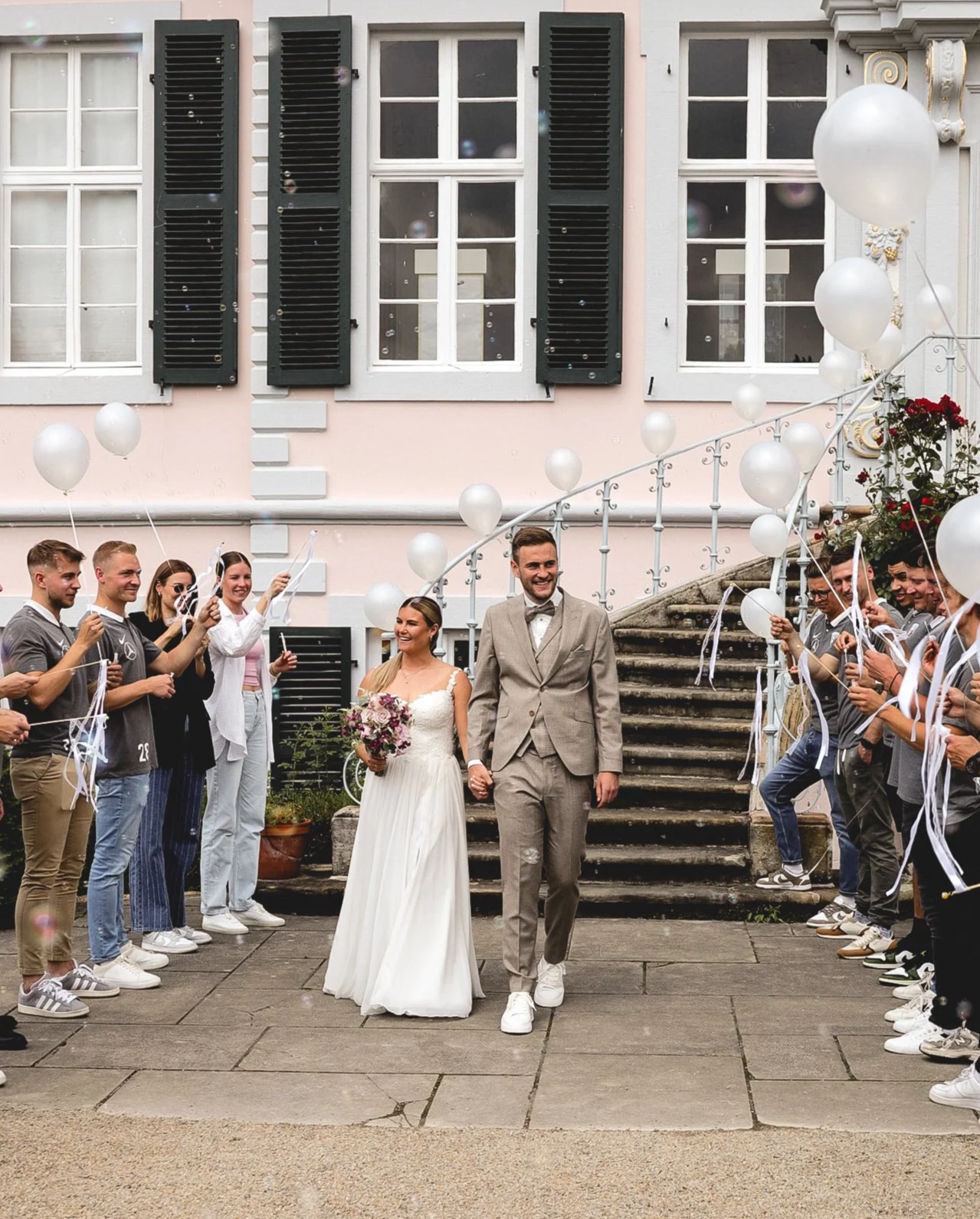 Ein Hochzeitspaar läuft Hand in Hand aus dem Standesamt. Die Gäste stehen Spalier. Im Hintergrund ein historisches Gebäude mit dunkelgrünen Fensterläden, am Treppengeländer sind Luftballon befestigt.