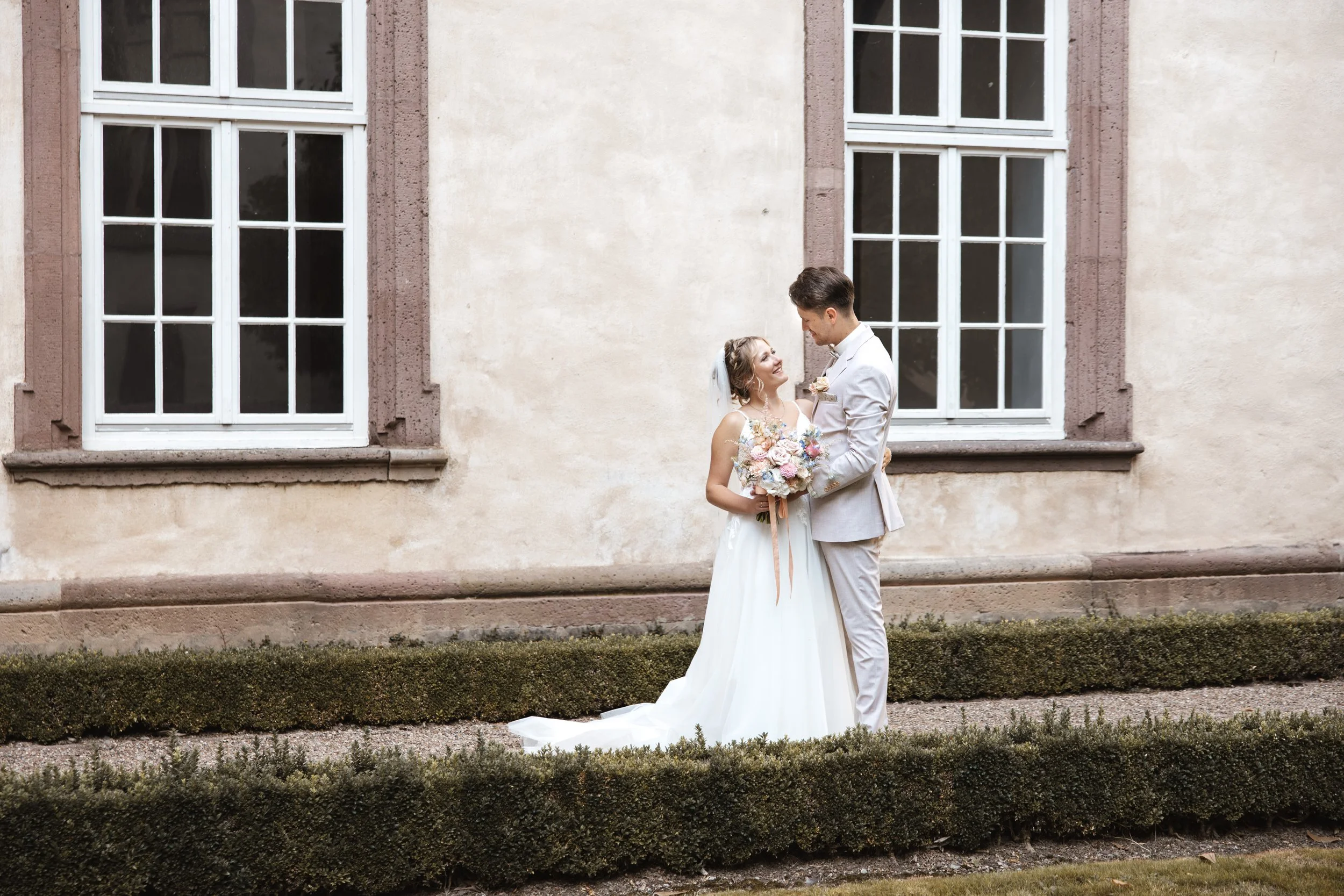 Ein Brautpaar steht vor einem alten Haus mit zwei großen Fenstern, die zu einem Hochzeitstag posieren.