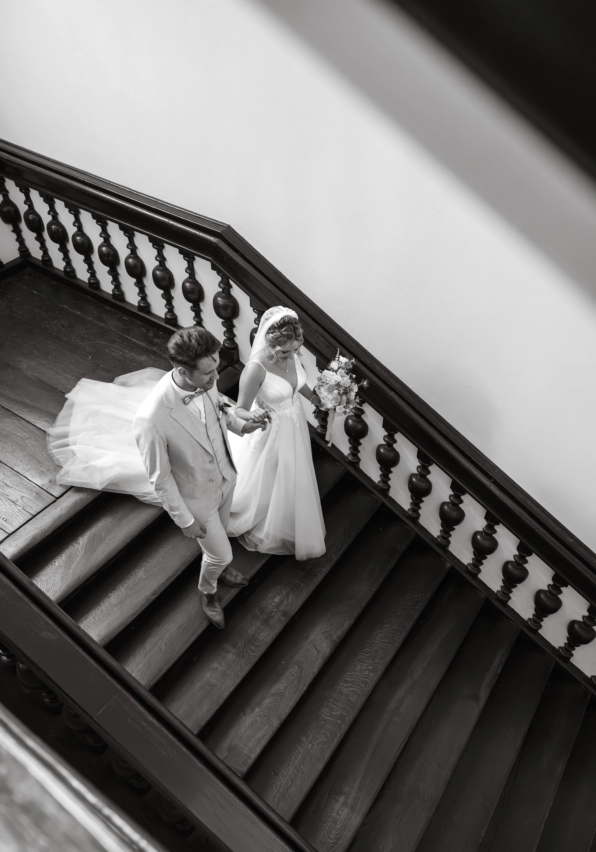 Schwarz-weiß-Foto eines Brautpaares, das die Treppe eines historischen Gebäudes hinuntergeht, die Braut trägt ein langes Kleid mit Schleier, der Bräutigam trägt einen hellen Anzug mit Krawatte.