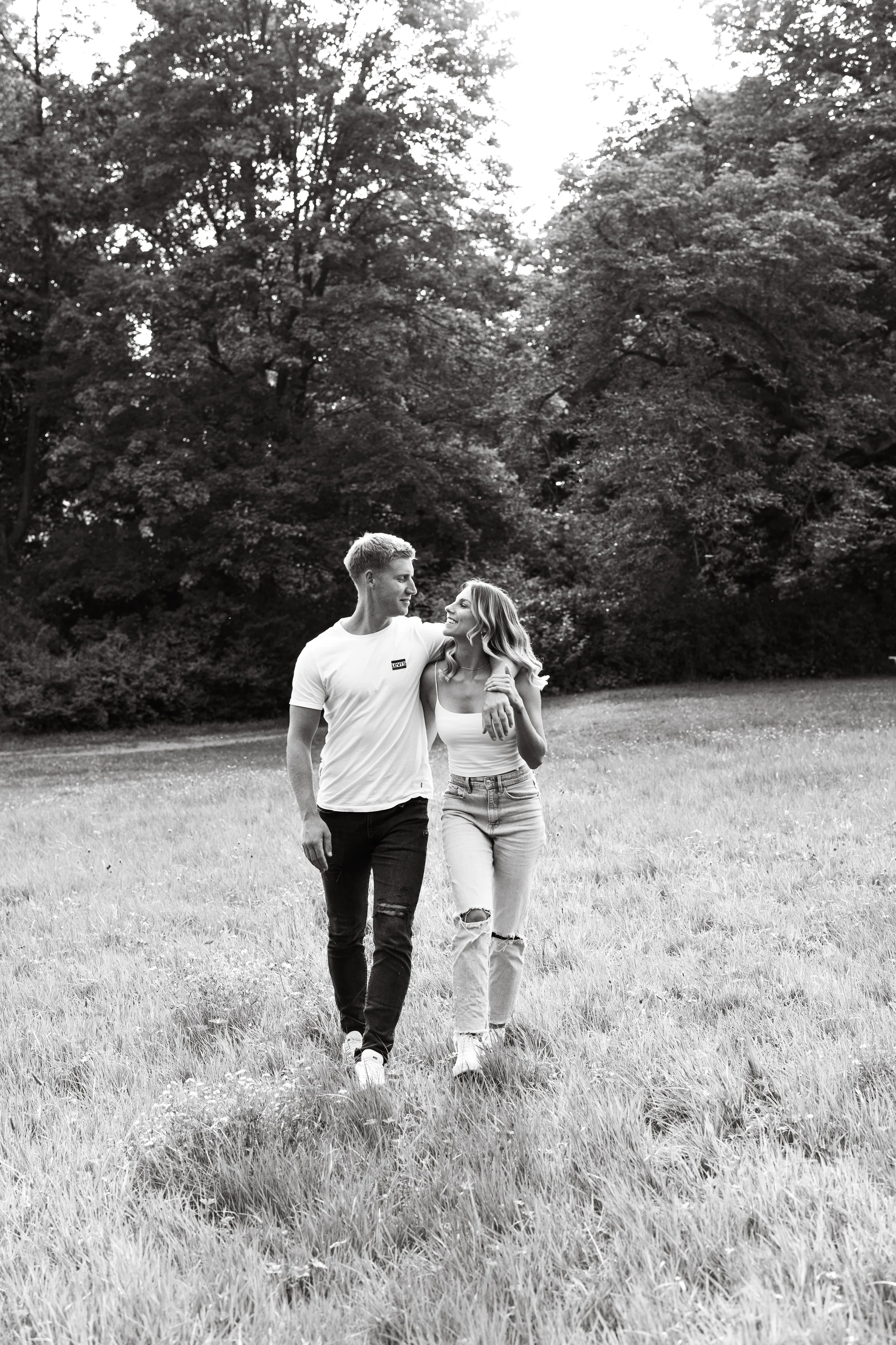 Ein junges Paar geht Arm in Arm durch eine Wiesenlandschaft mit Bäumen im Hintergrund, Schwarz-Weiß-Fotografie.