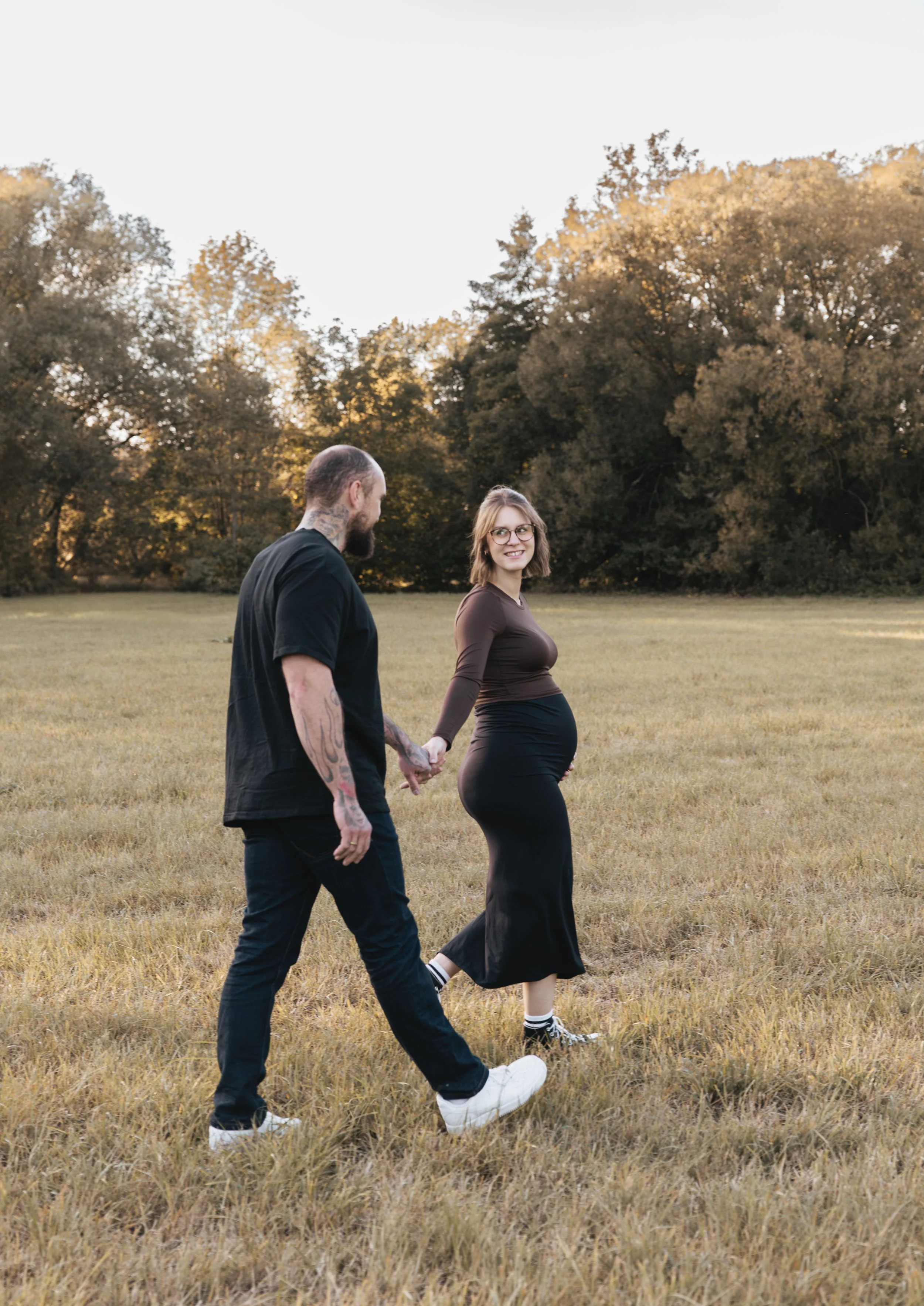 Ein schwangeres Paar läuft Hand in Hand über eine offene, grüne Wiese.