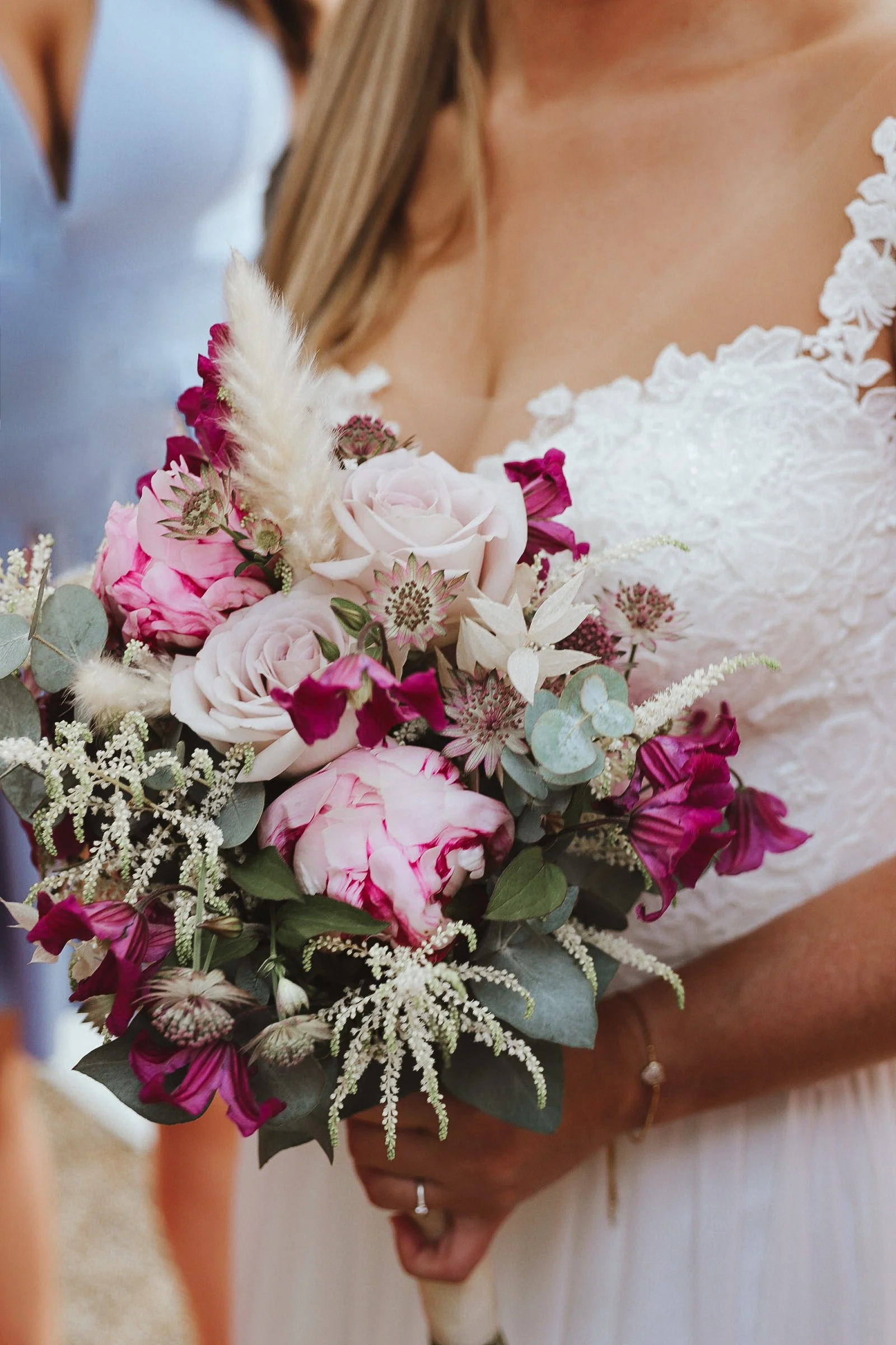 Nahaufnahme eines Brautstraußes mit rosa und weißen Blumen, grünen Blättern und dekorativen weißen Federn, gehalten von einer Braut in einem weißen Spitzenkleid.