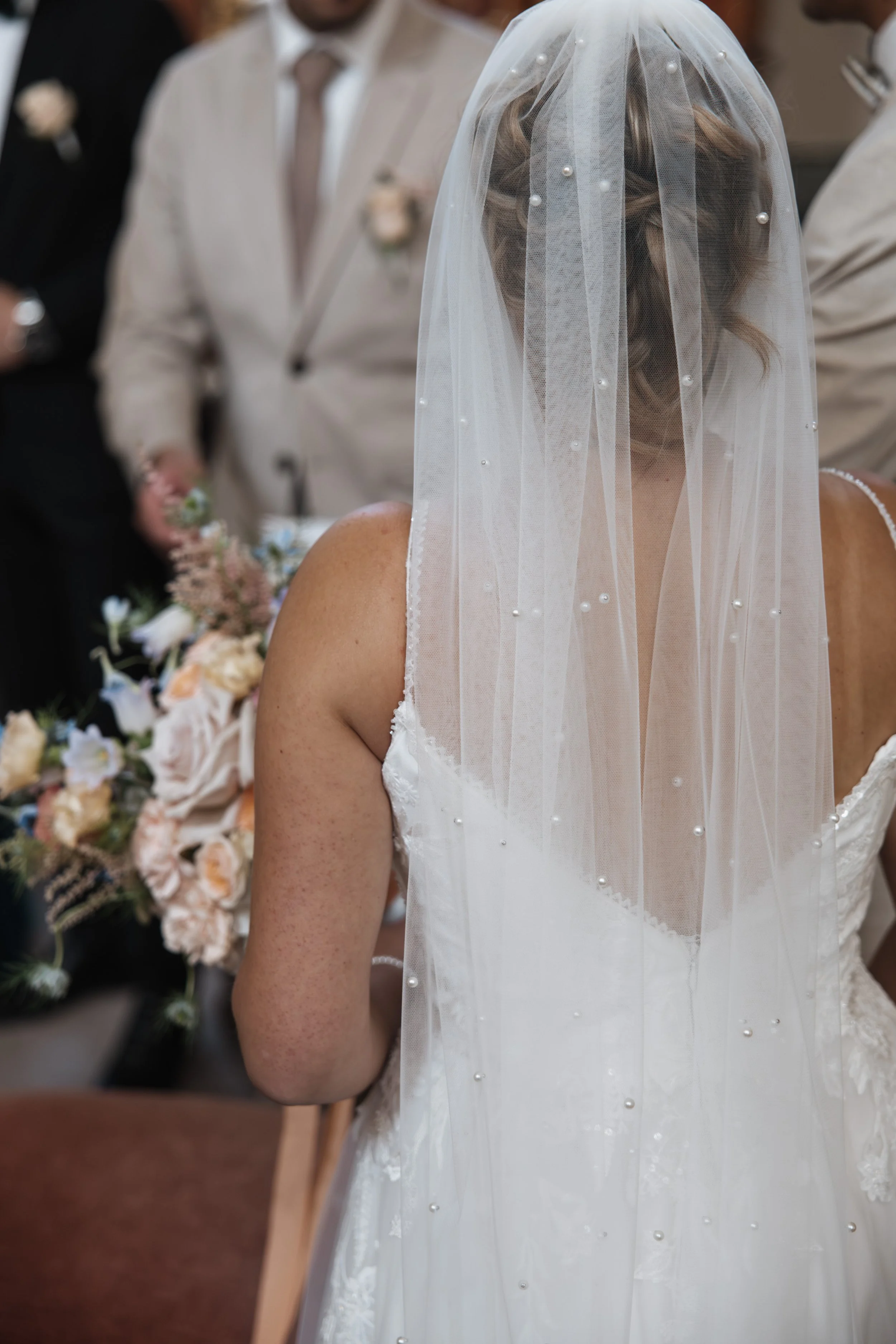 Braut mit weißem Kleid und Schleier, umgeben von Gästen bei einer Hochzeit.