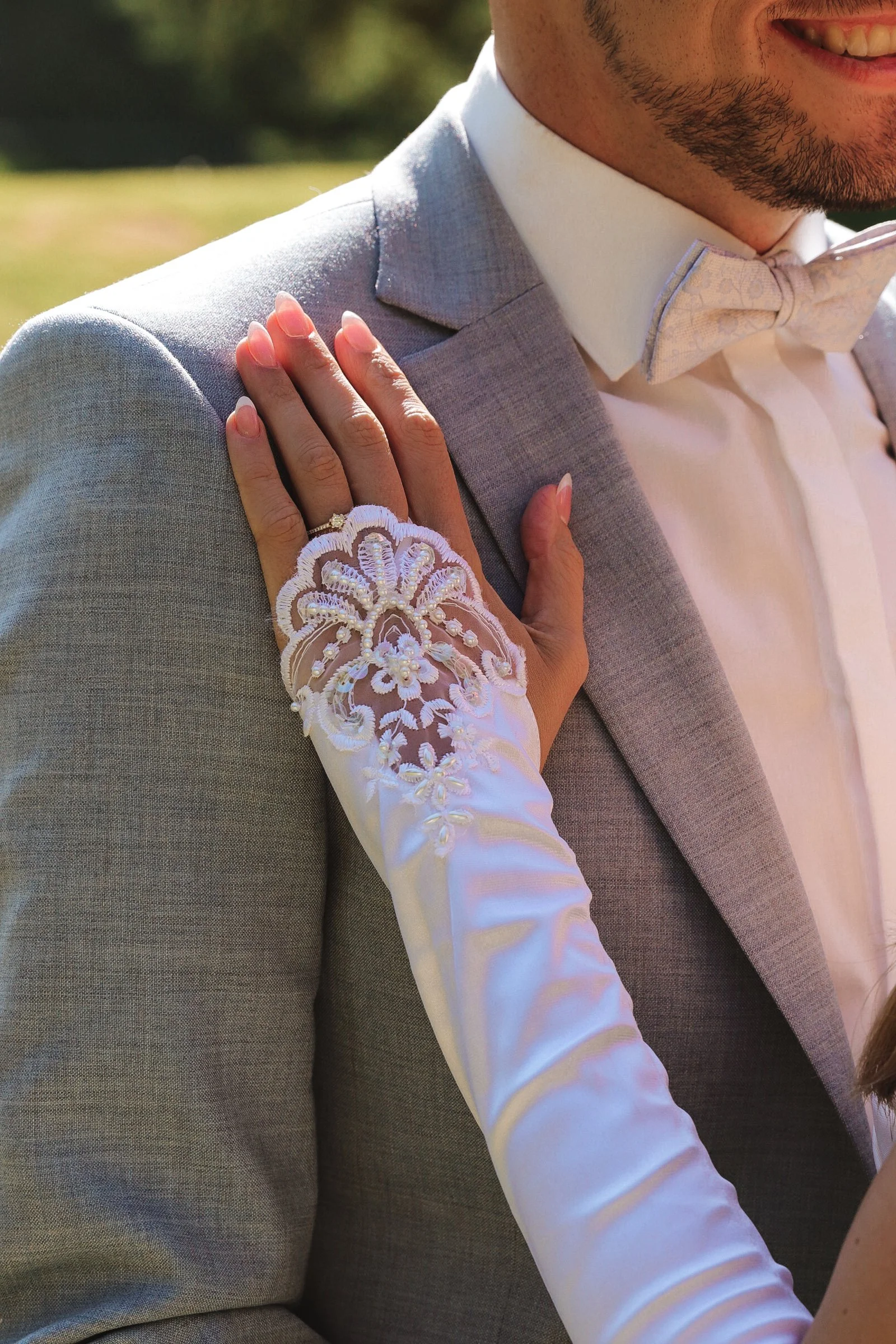 Nahaufnahme eines Brautpaares, bei der die Braut ihre Hand auf die Schulter des Bräutigams legt. Der Bräutigam trägt einen grauen Anzug und eine weiße Fliege. Sie trägt einen weißen Spitzenhandschuh und ihren Verlobungsring.