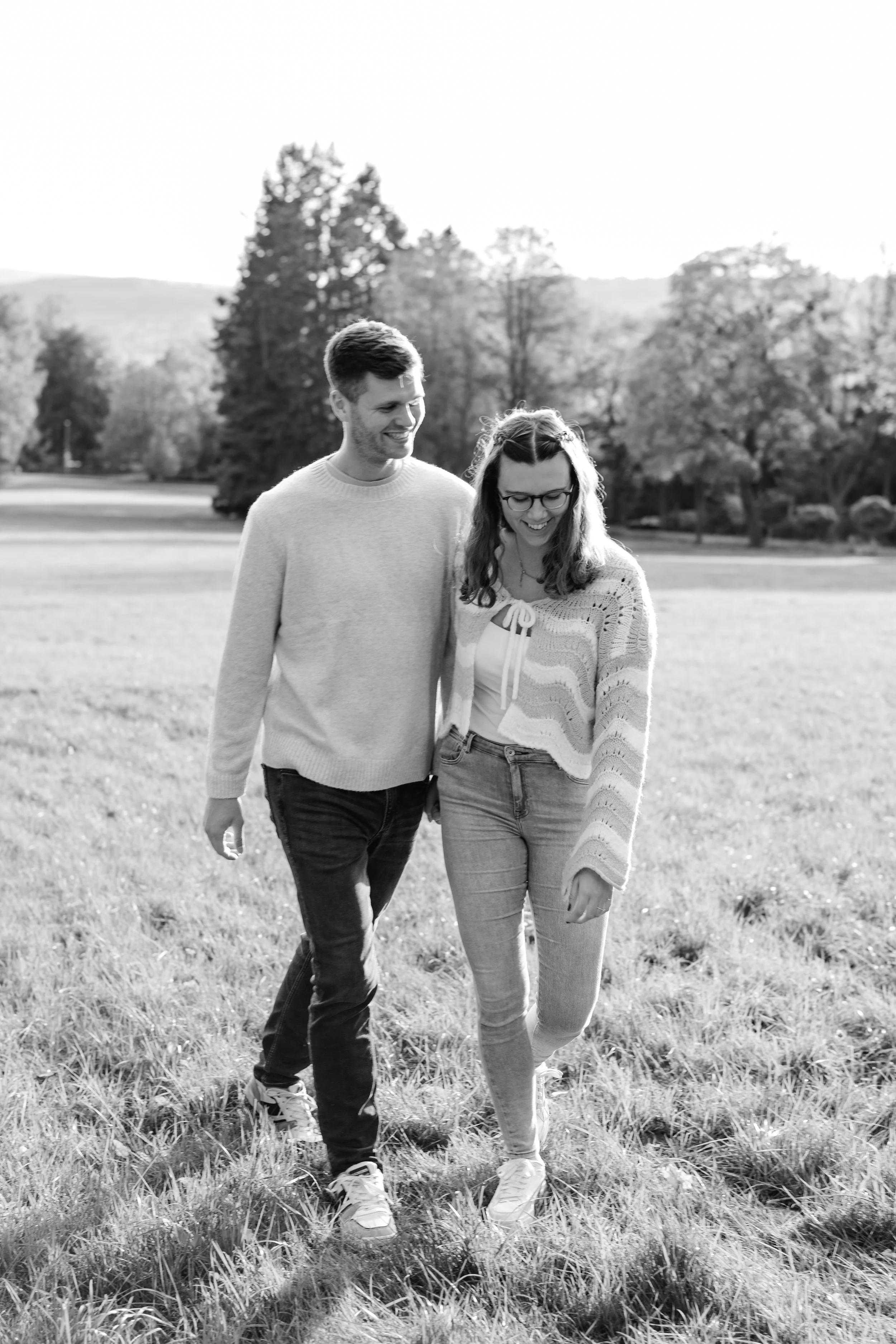 Ein lachendes Paar spaziert gemeinsam durch ein offenes grünes Feld mit Bäumen im Hintergrund. Schwarz-weiß Foto.