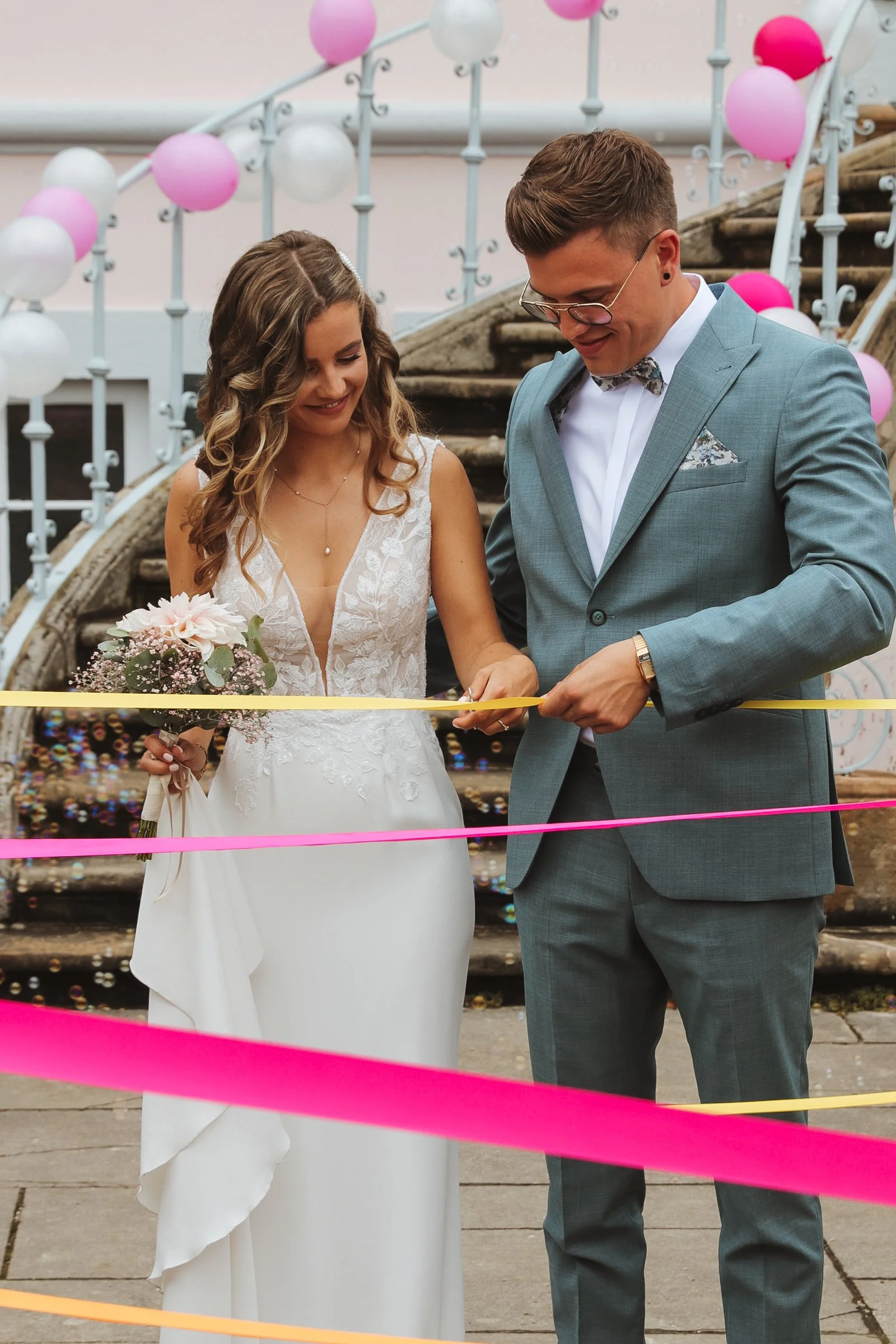 Ein Brautpaar bei der Hochzeitszeremonie, das zusammen bunte Schleifne durchschneidet, beim Auszug aus dem Standesamt. 