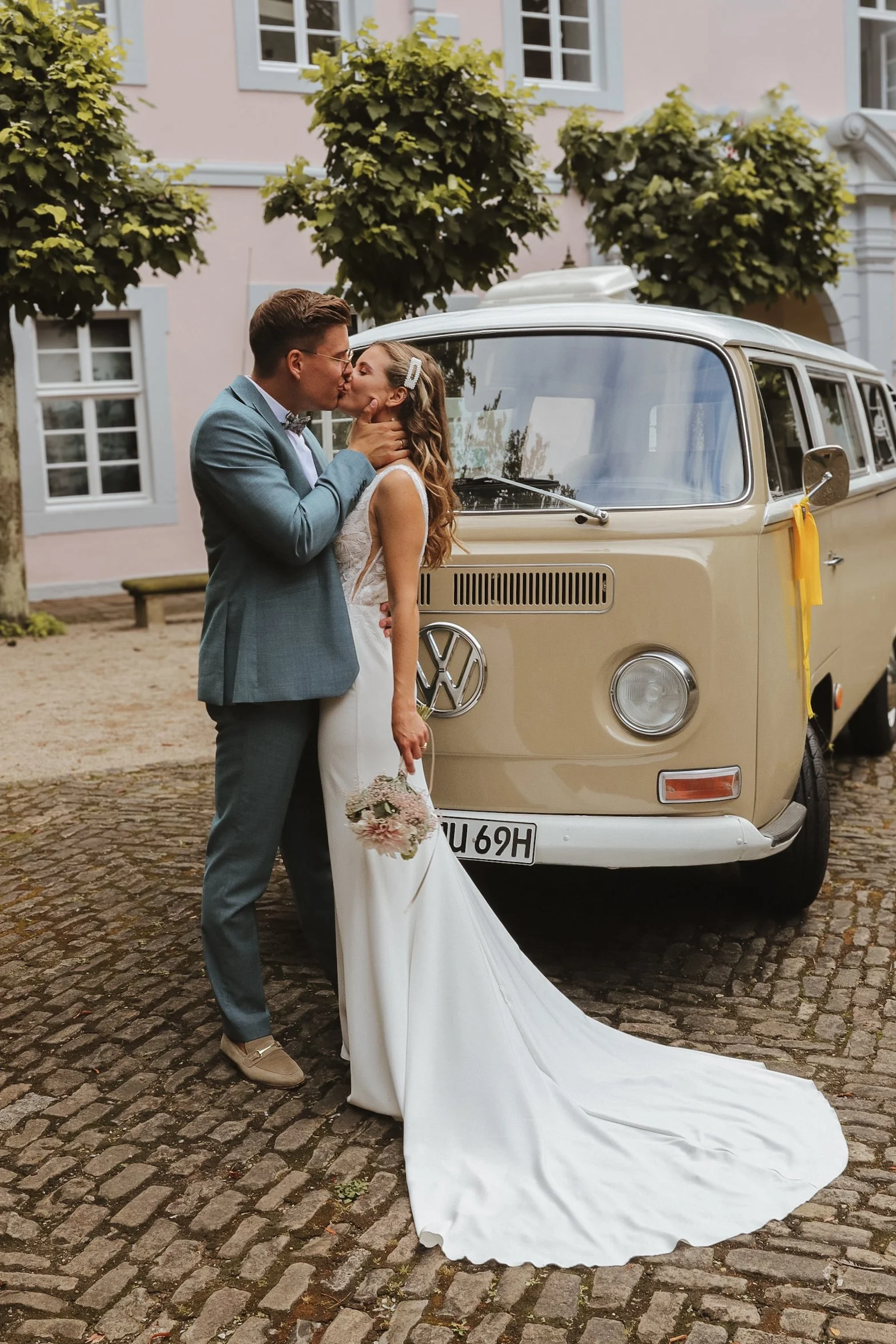 Ein Ehepaar küsst sich vor einem vintage VW Bus, umgeben von Bäumen und historischen Gebäuden. Sie trägt ein langes Brautkleid mit Schleppe, er einen blauen Anzug. 