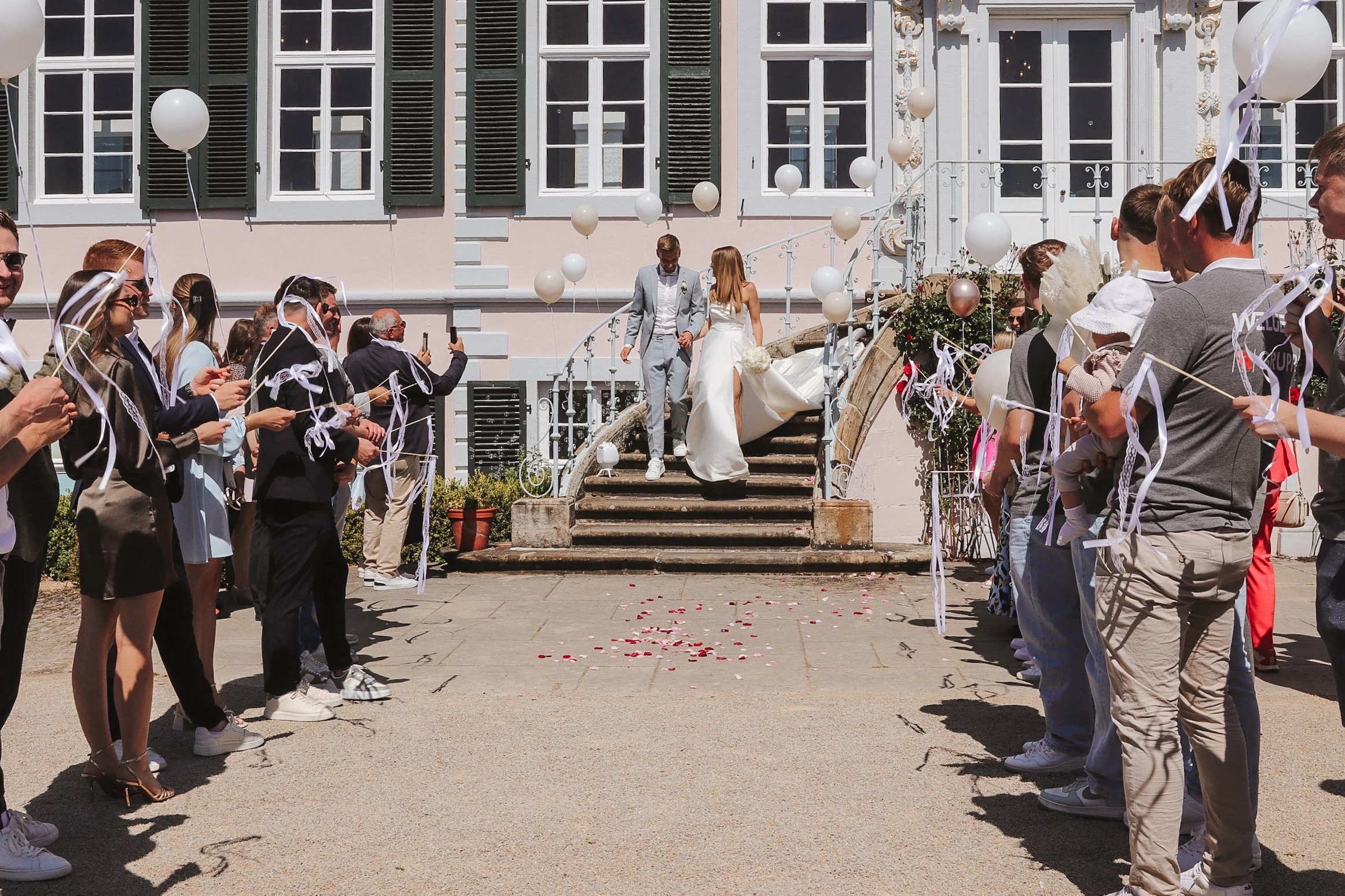 Brautpaar auf Stufen vor einem historischen Haus, umgeben von Gästen, die Luftballons halten, beim Auszug aus dem Standesamt.