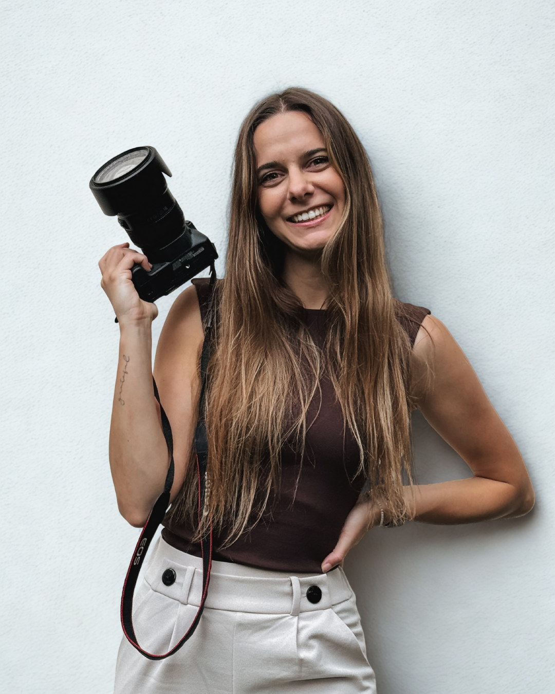 Porträt einer jungen Fotografin mit langen, glatten brünetten Haaren. Sie lächelt freundlich in die Kamera und hält ihre Kamera in ihrer rechten Hand nach oben. Linker Arm ist angewinkelt und in ihre Taille gestemmt.