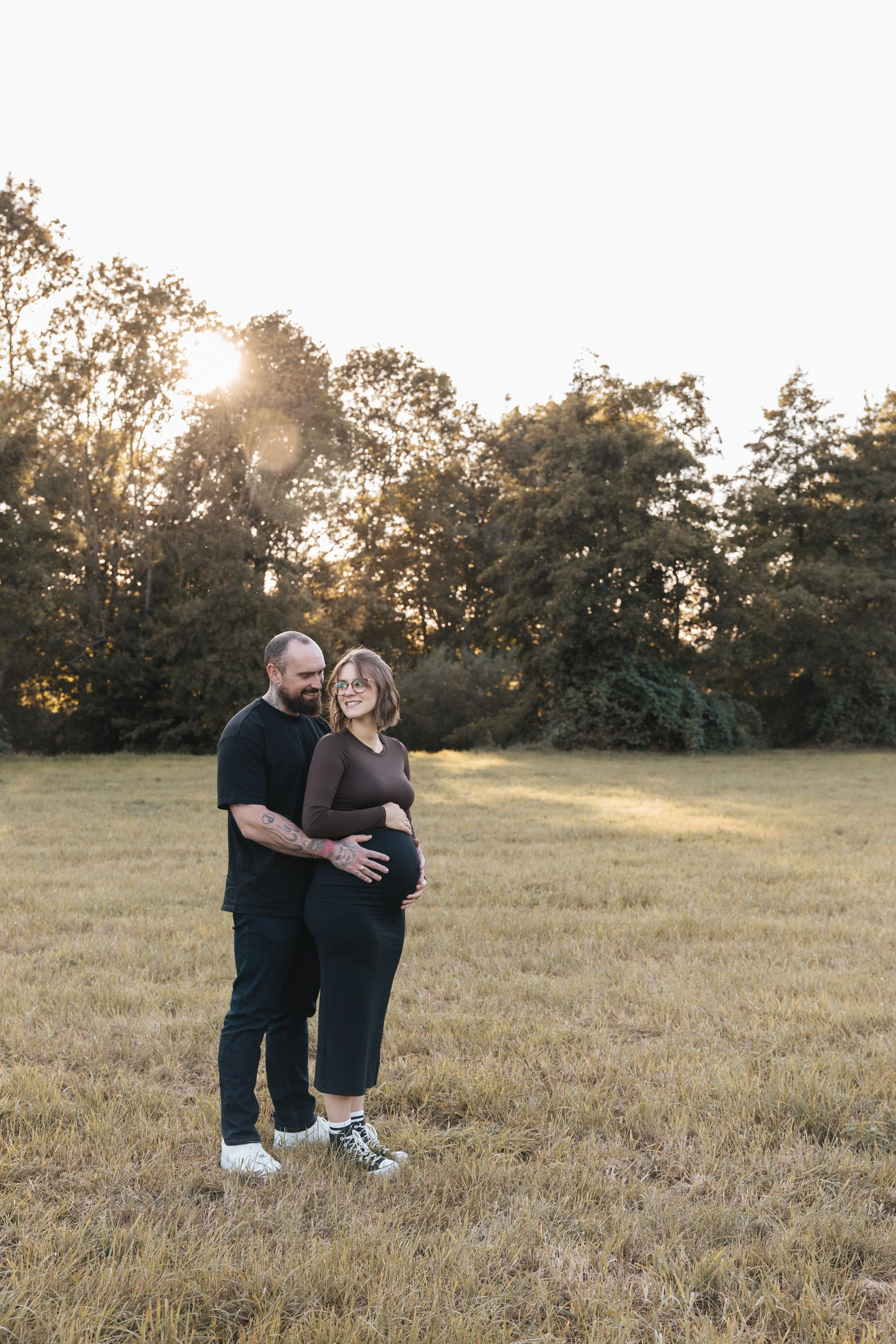 Ein schwangeres Paar steht auf einer Wiese bei Sonnenuntergang und lächelt. Die Frau trägt dunkle Kleidung, während der Mann schwarze Kleidung trägt und Tattoos hat.