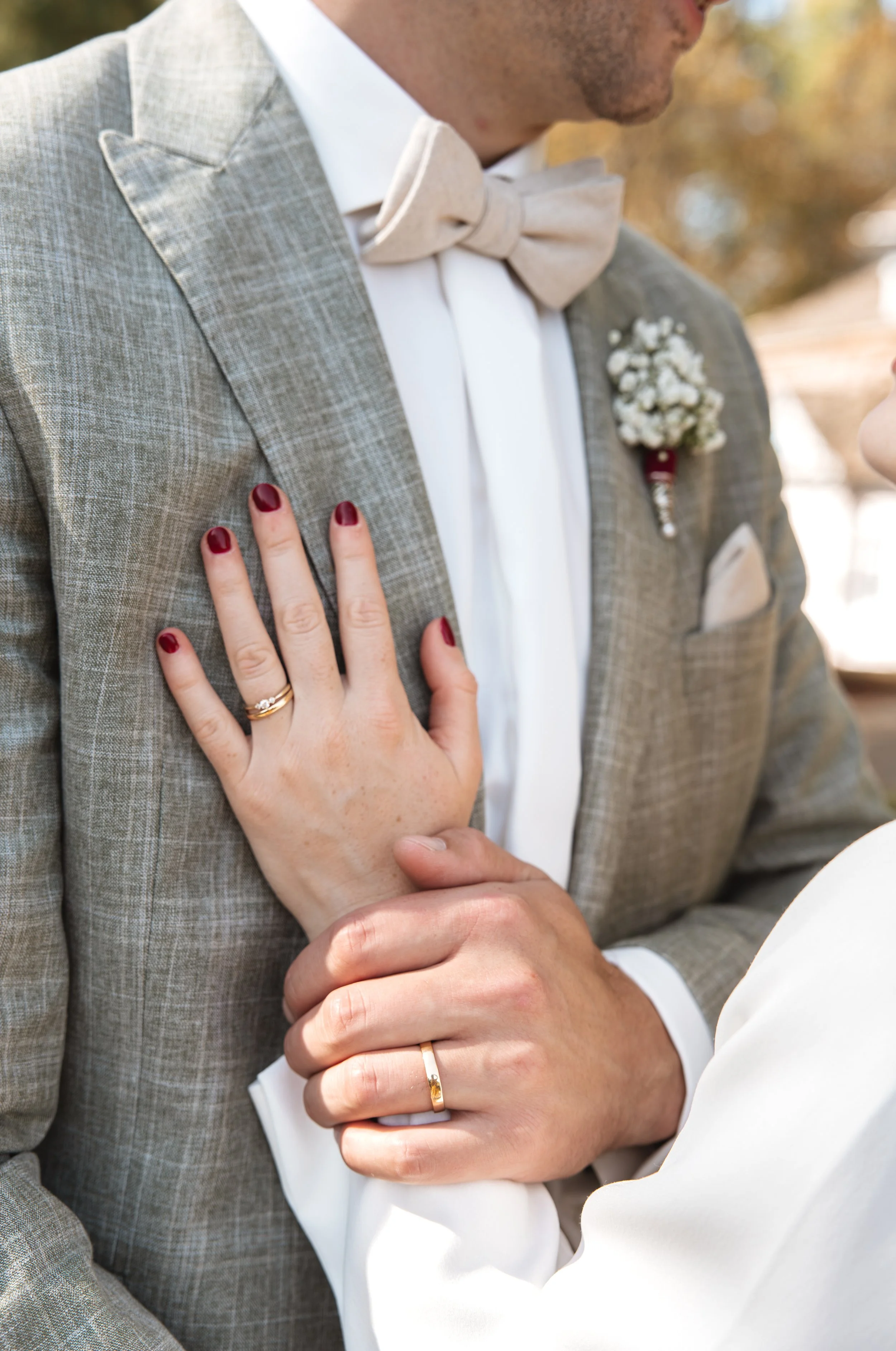 Ein Mann und eine Frau, die sich bei einer Hochzeit küssen, sehen sich liebevoll an. Der Mann trägt einen beigen Anzug mit Fliege, die Frau trägt einen weißen Mantel und rote Nagellack. Beide tragen Eheringe.