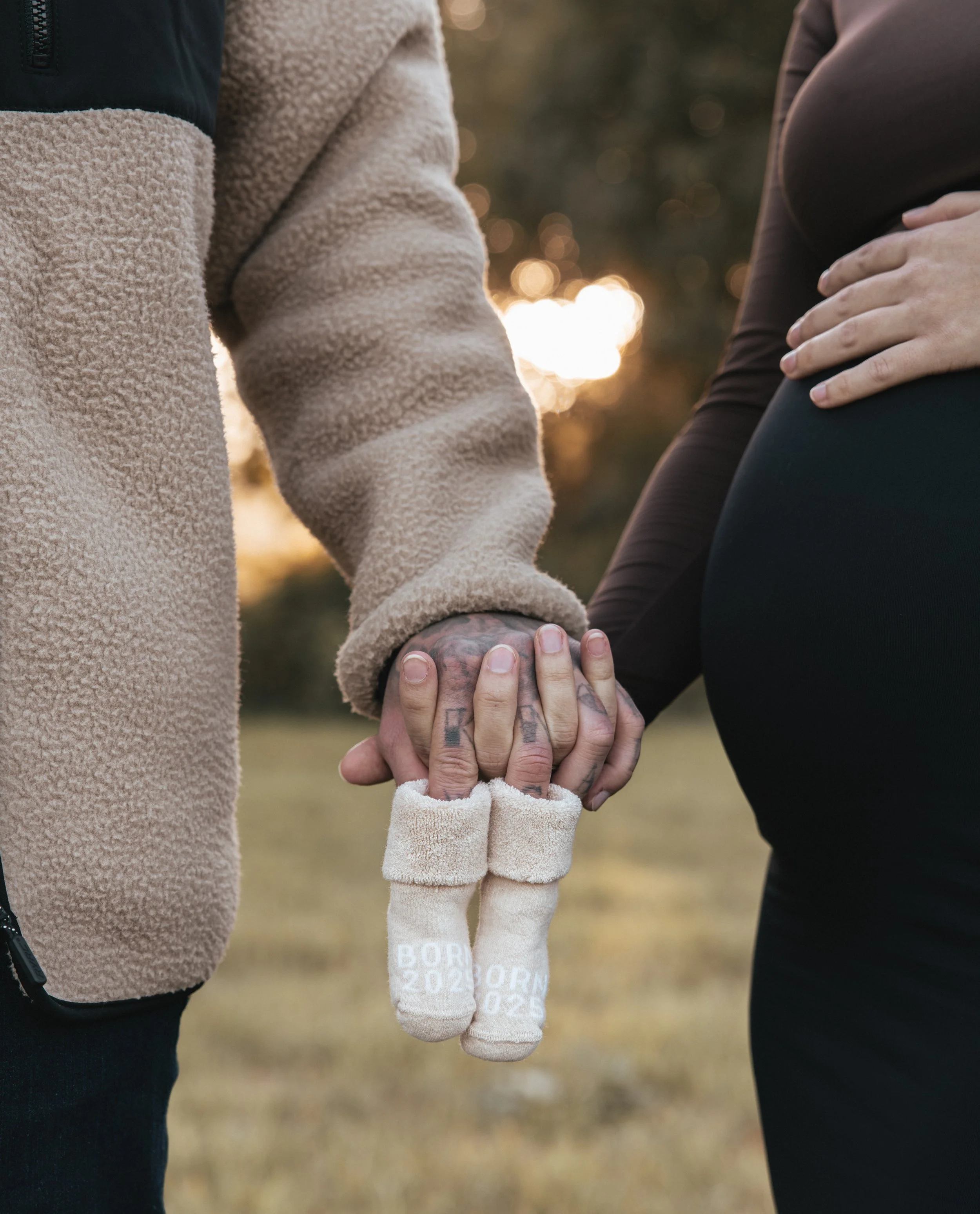 Ein Paar Hände hält sich im Freien bei Sonnenuntergang Händchen, während eine schwangere Frau ihre Hand auf ihren Bauch legt. In der Hand halten sie kleine Babysöckchen.