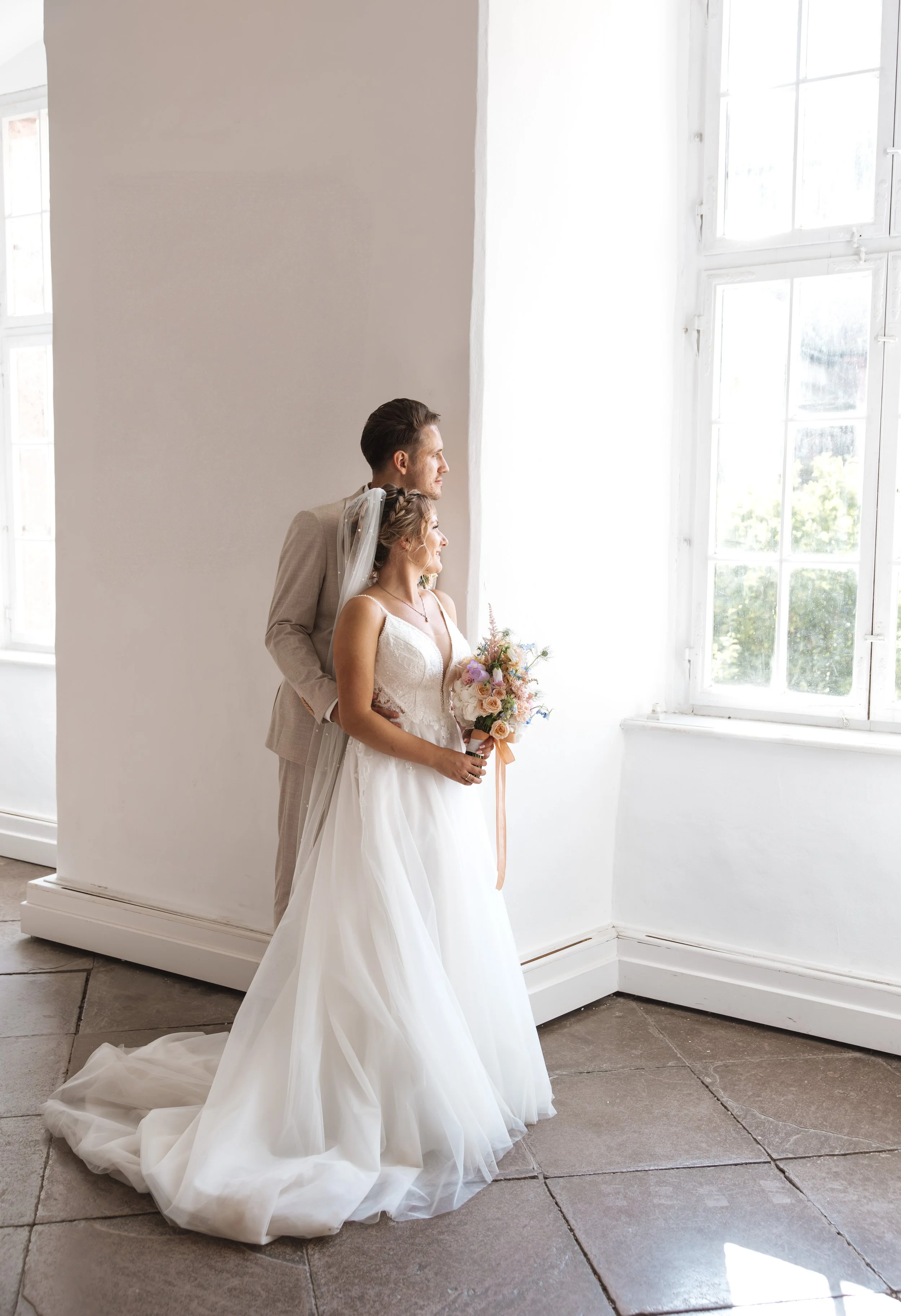 Ein Brautpaar steht gemeinsam vor einem großen Fenster in einem hellen Raum, der Bräutigam trägt einen hellen Anzug, die Braut hält einen Blumenstrauß und trägt ein weißes Hochzeitskleid.