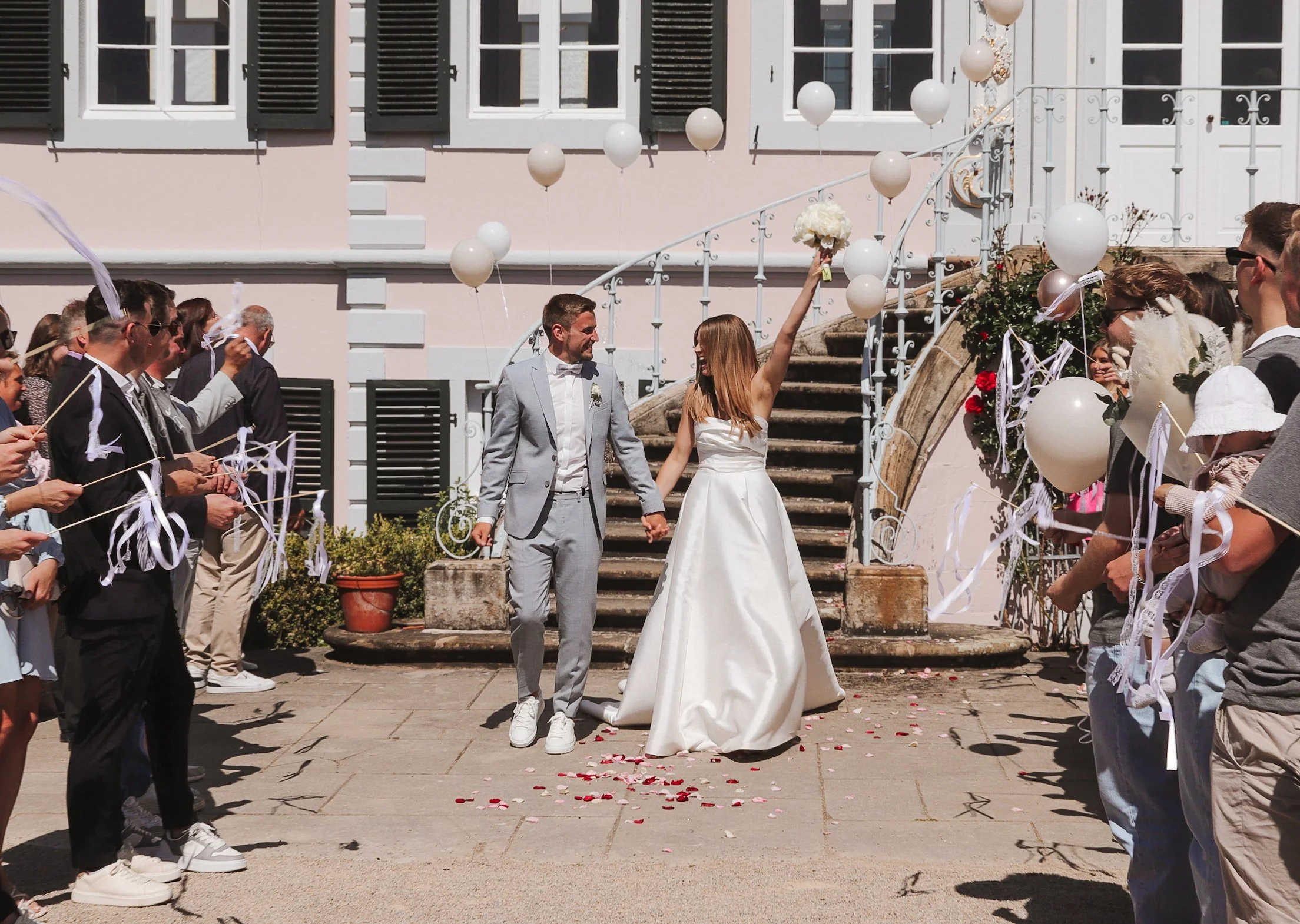 Das Brautpaar beim Auszug aus dem Standesamt, gehen durch ein Spalier der Gäste. Das Brautpaar freut sich, die Braut hebt einen Brautstrauß in die Luft. Die Gäste werfen Blumenblätter. Es ist ein sonniger Tag.