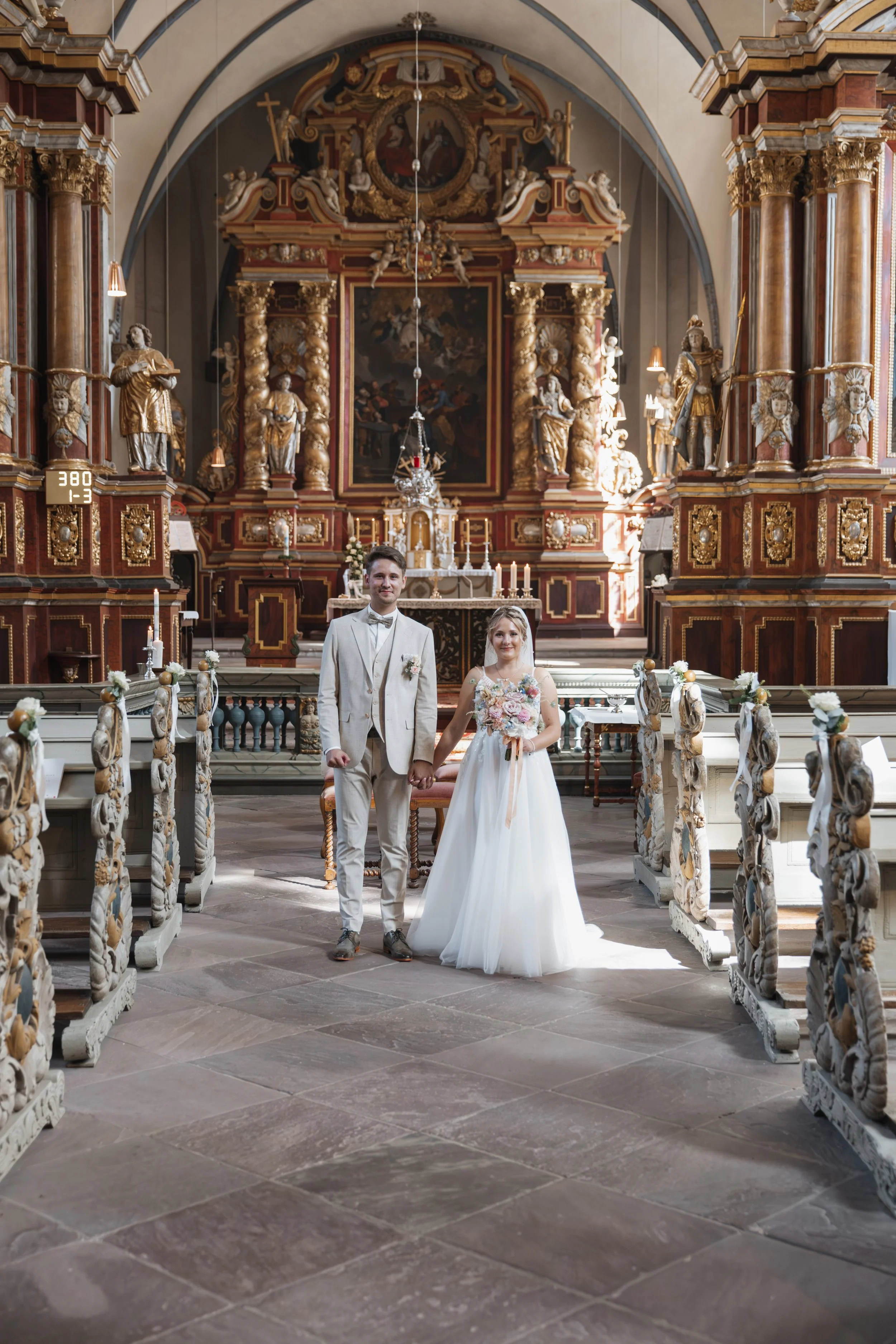 Ein Brautpaar steht in einer Kirche. Der Bräutigam trägt einen hellen Anzug, die Braut trägt ein weißes Hochzeitskleid und hält einen Blumenstrauß. Die Kirche ist reich verziert mit Holzschnitzereien und Barock-Details.