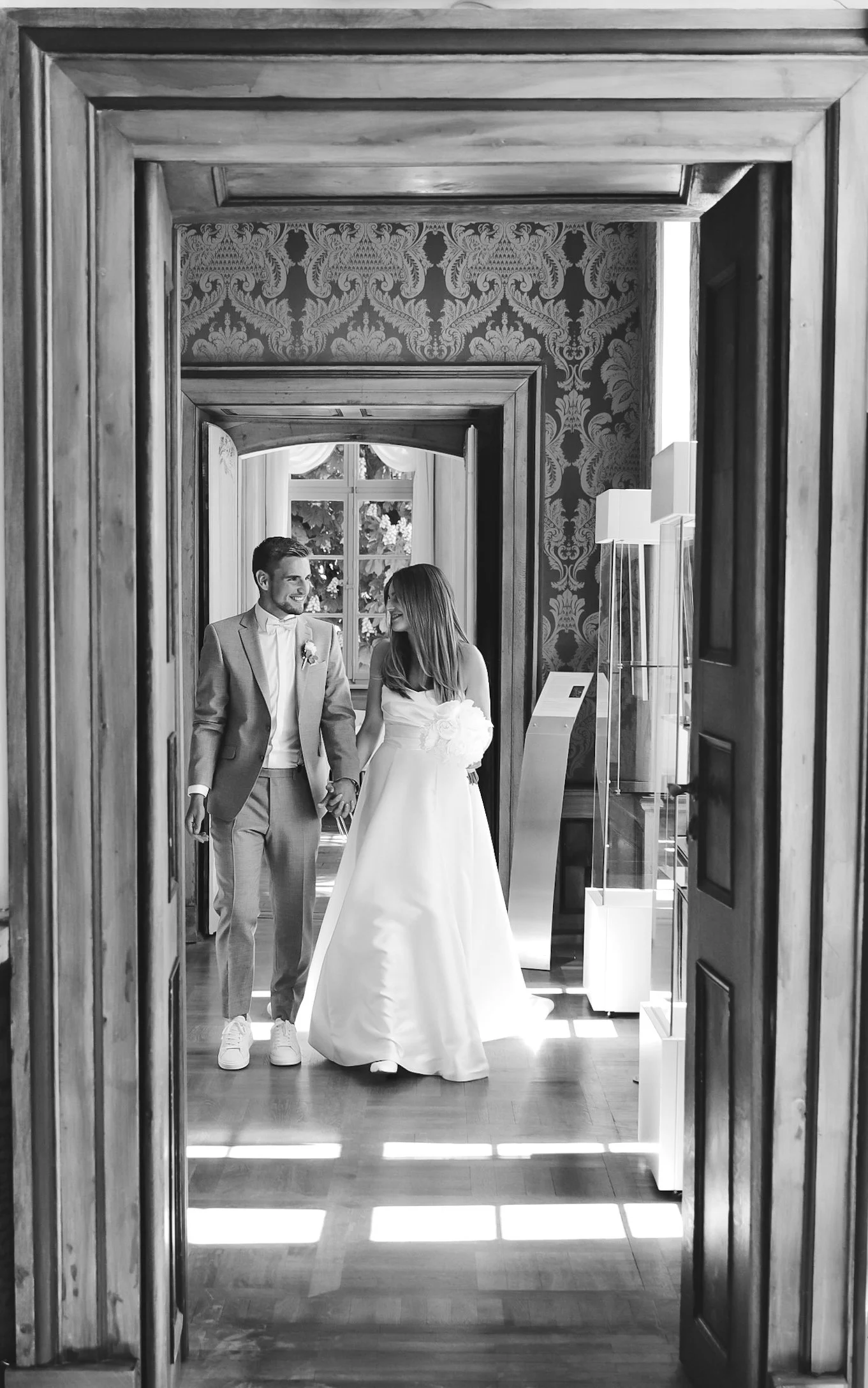 Zwei Menschen, ein Mann in einem Anzug und eine Frau in einem Brautkleid, die Händchen halten und sich ansehen, durch einen offenen Holztürrahmen in einen eleganten Raum mit dekorativer Tapete und großen Fensters gehen. Schwarz-Weiß-Fotografie.