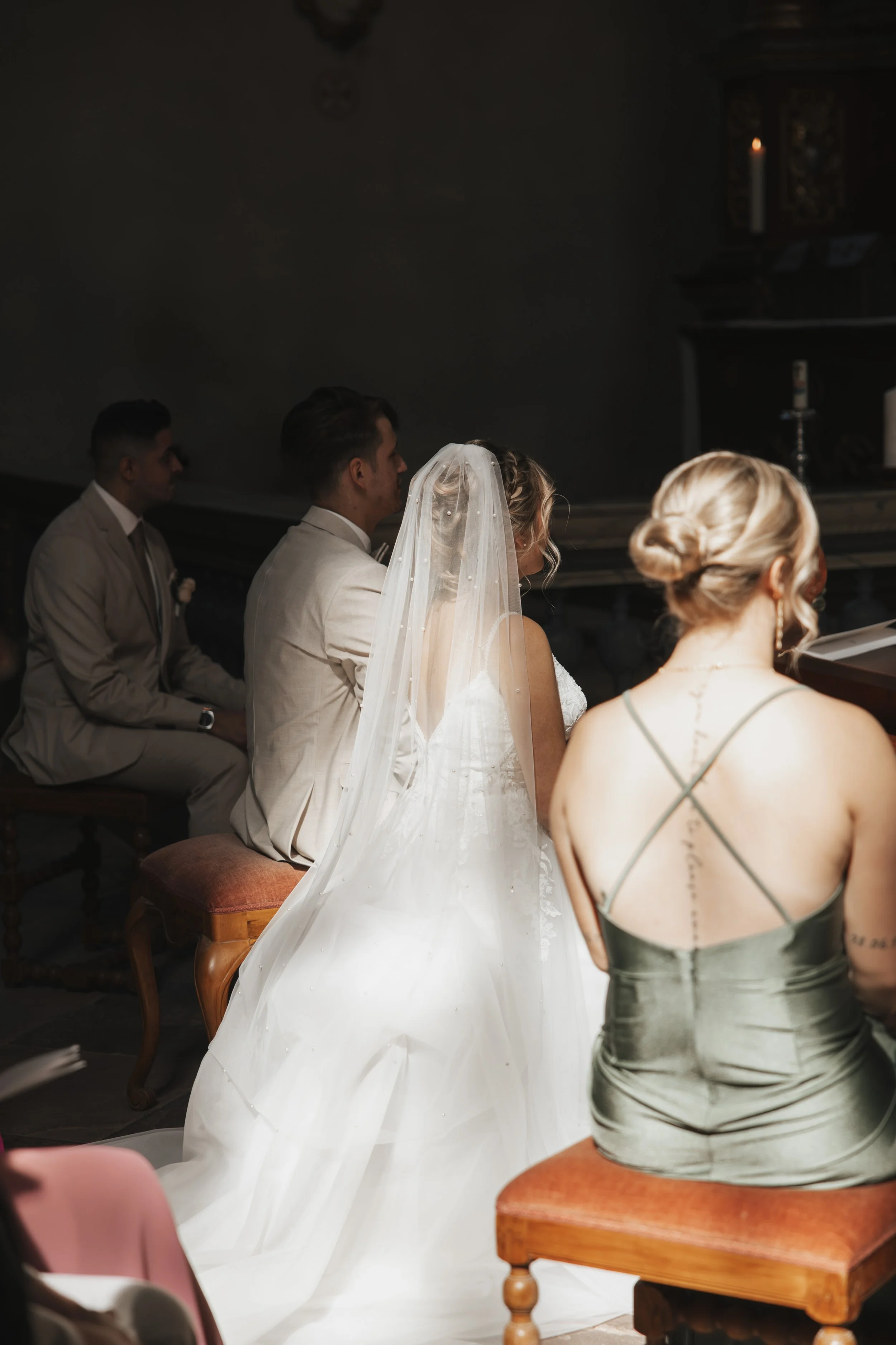 Braut in Weißer Hochzeitskleidung mit Schleier, sitzend bei einer Zeremonie, neben einer Frau in grünem Kleid, im Hintergrund Männer in Anzügen