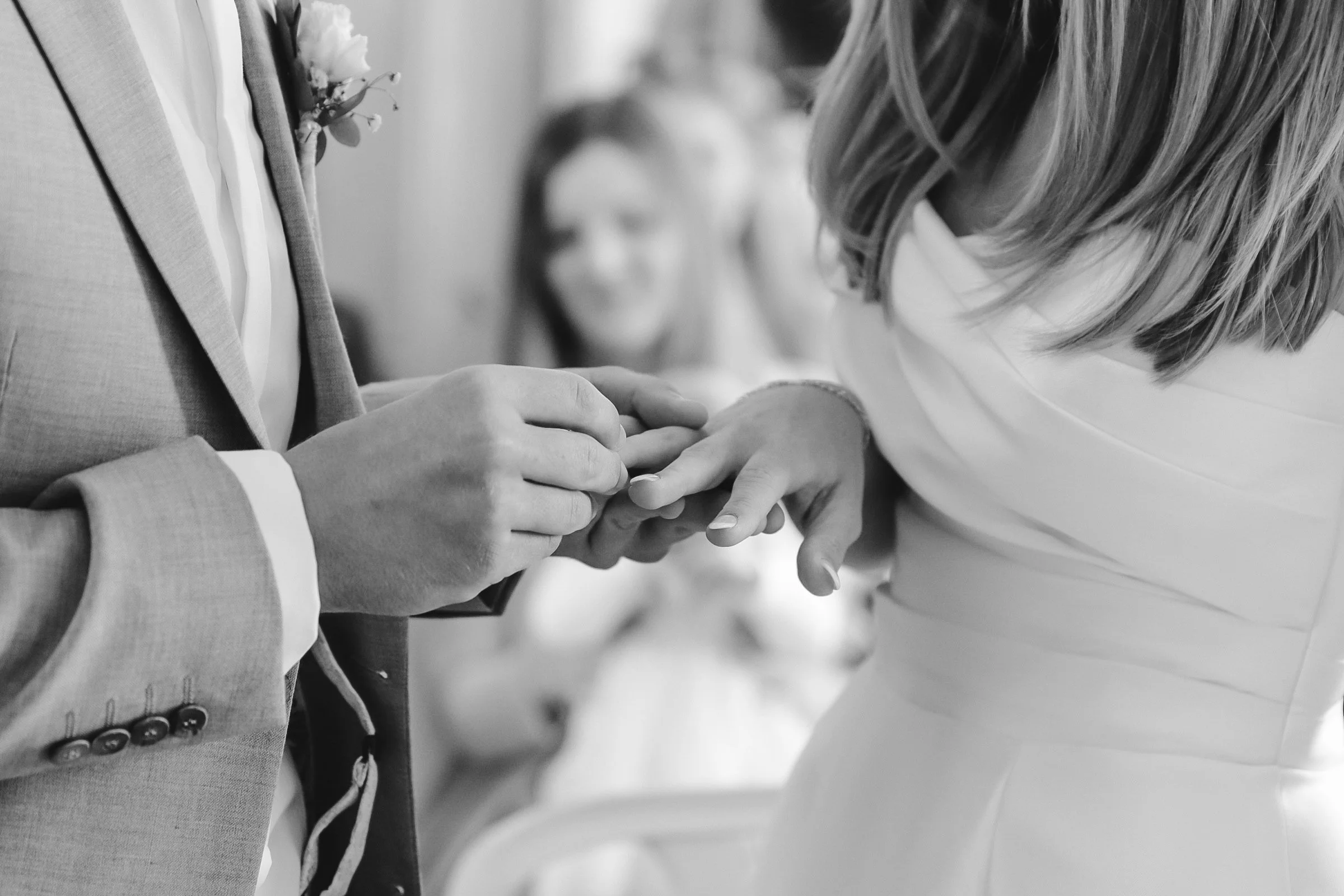 Ringtausch während der standesamtlichen Trauung. Der Mann legt der Frau einen Ring an den Finger, im Schwarz-Weiß-Foto, mit einer lachenden Frau im Hintergrund.